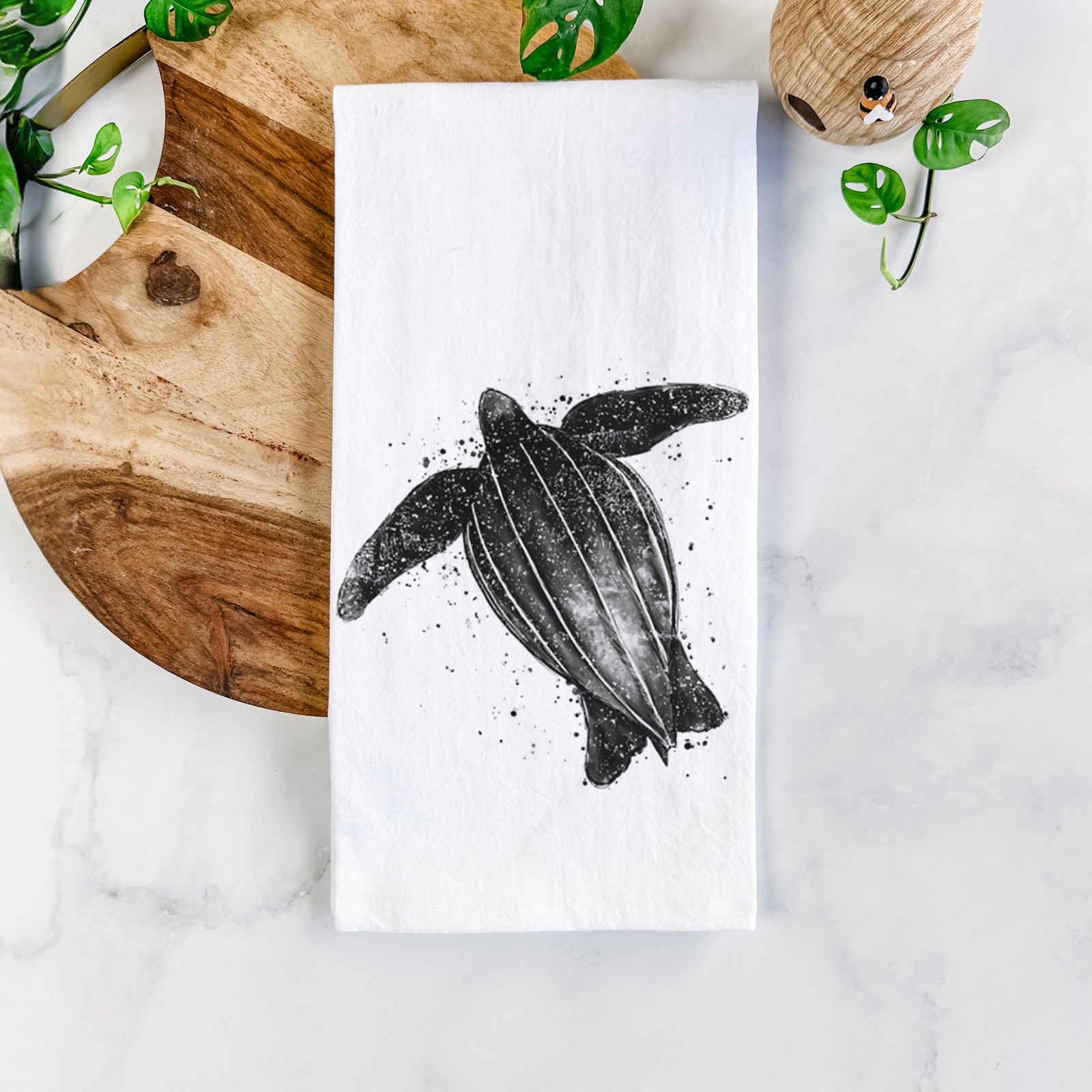 White cotton tea towel printed with a black ink illustration of a leatherback sea turtle swimming, with ink splatters, on a marble counter beside a round wooden cutting board and greenery.