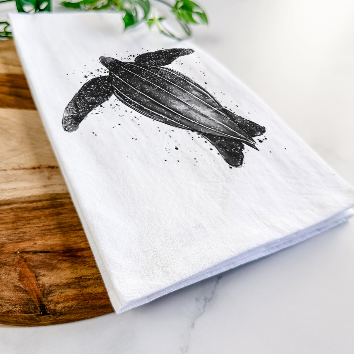 Close-up of a folded white tea towel on a wooden board, showing a black ink illustration of a leatherback sea turtle swimming, with ink splatters.