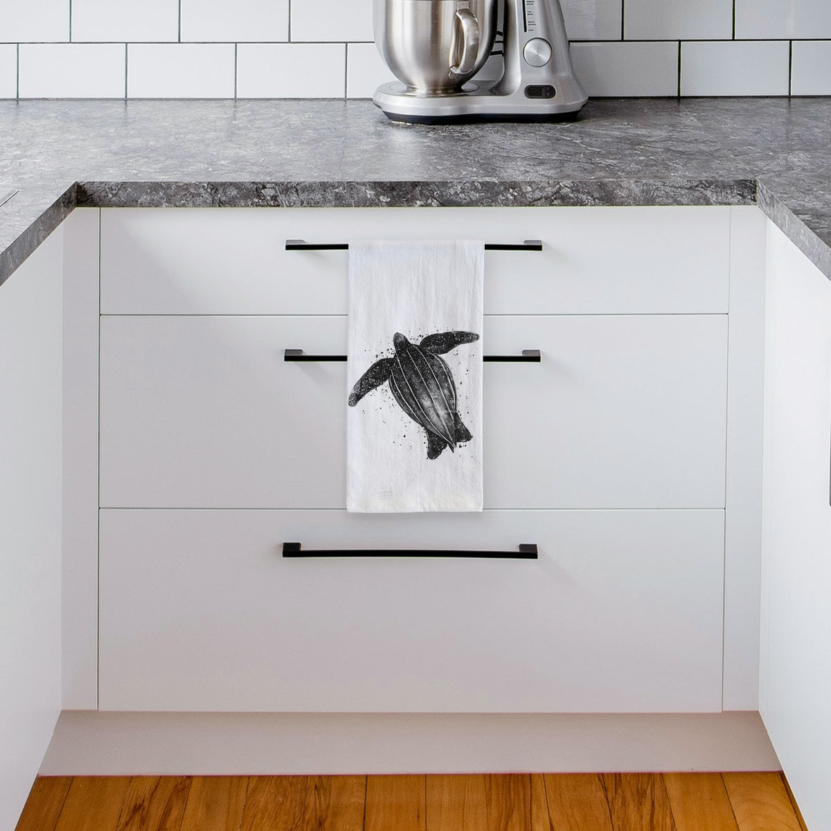 White tea towel printed with a black ink illustration of a leatherback sea turtle swimming, with ink splatters, hanging from a drawer handle in a bright white kitchen.