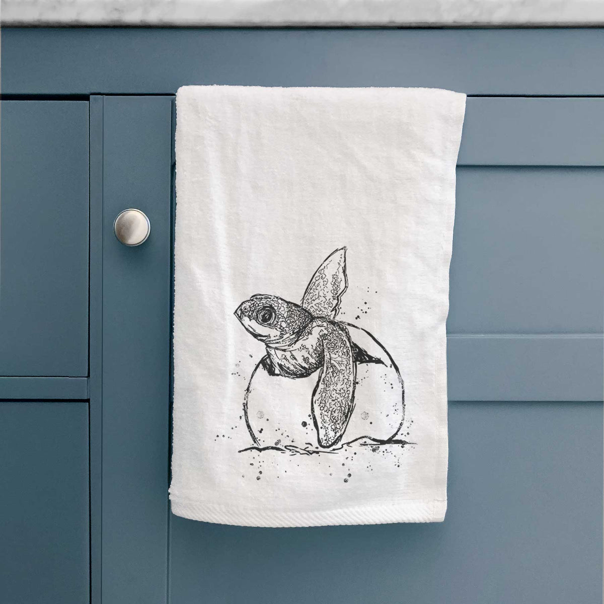 White hand towel printed with a black ink illustration of a leatherback sea turtle hatchling emerging from its egg, hanging on a cabinet door.