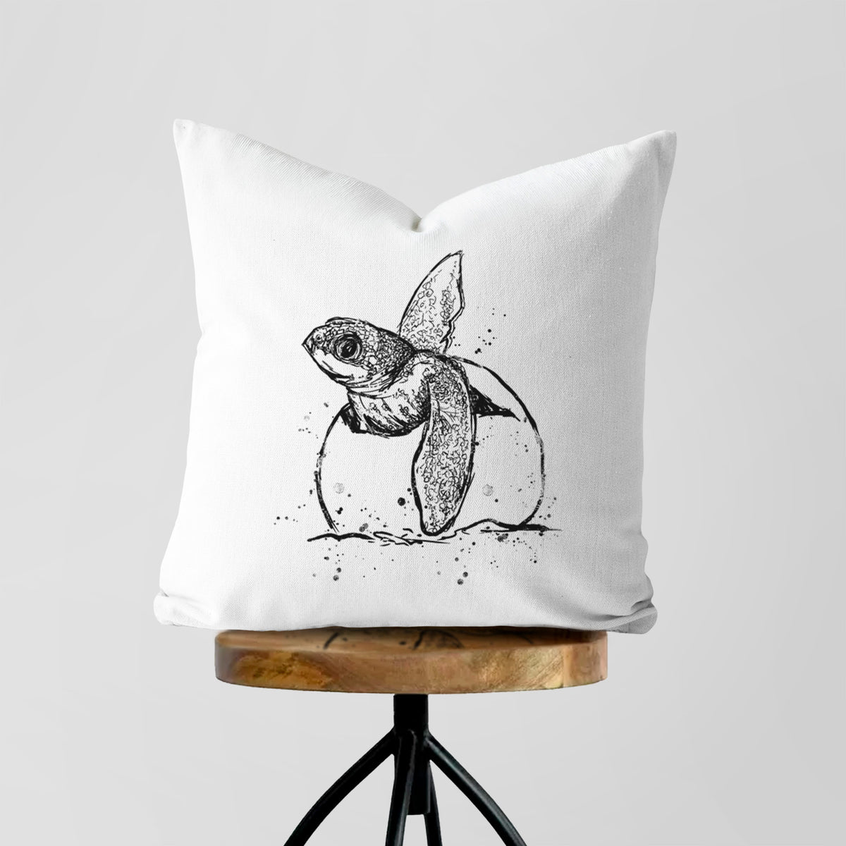 White pillow cover printed with a black ink illustration of a leatherback sea turtle hatchling emerging from its egg, on a wooden stool.