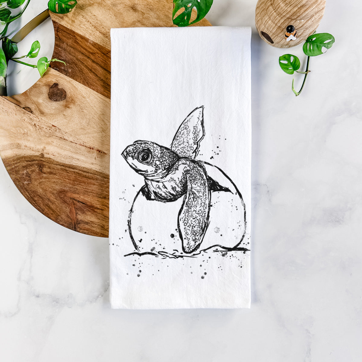 White cotton tea towel printed with a black ink illustration of a leatherback sea turtle hatchling emerging from its egg, on a marble counter beside a round wooden cutting board and greenery.