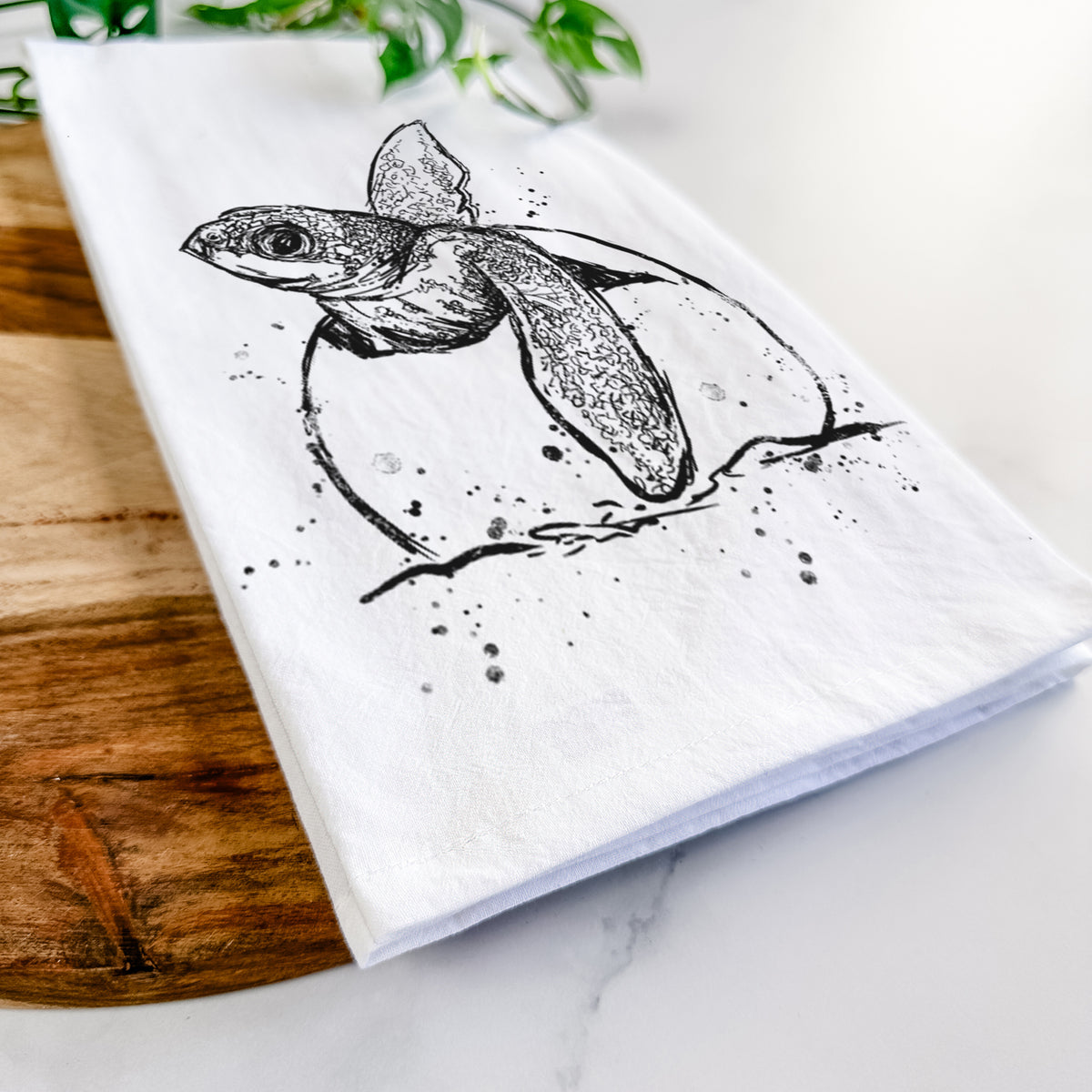 Close-up of a folded white tea towel on a wooden board, showing a black ink illustration of a leatherback sea turtle hatchling emerging from its egg.