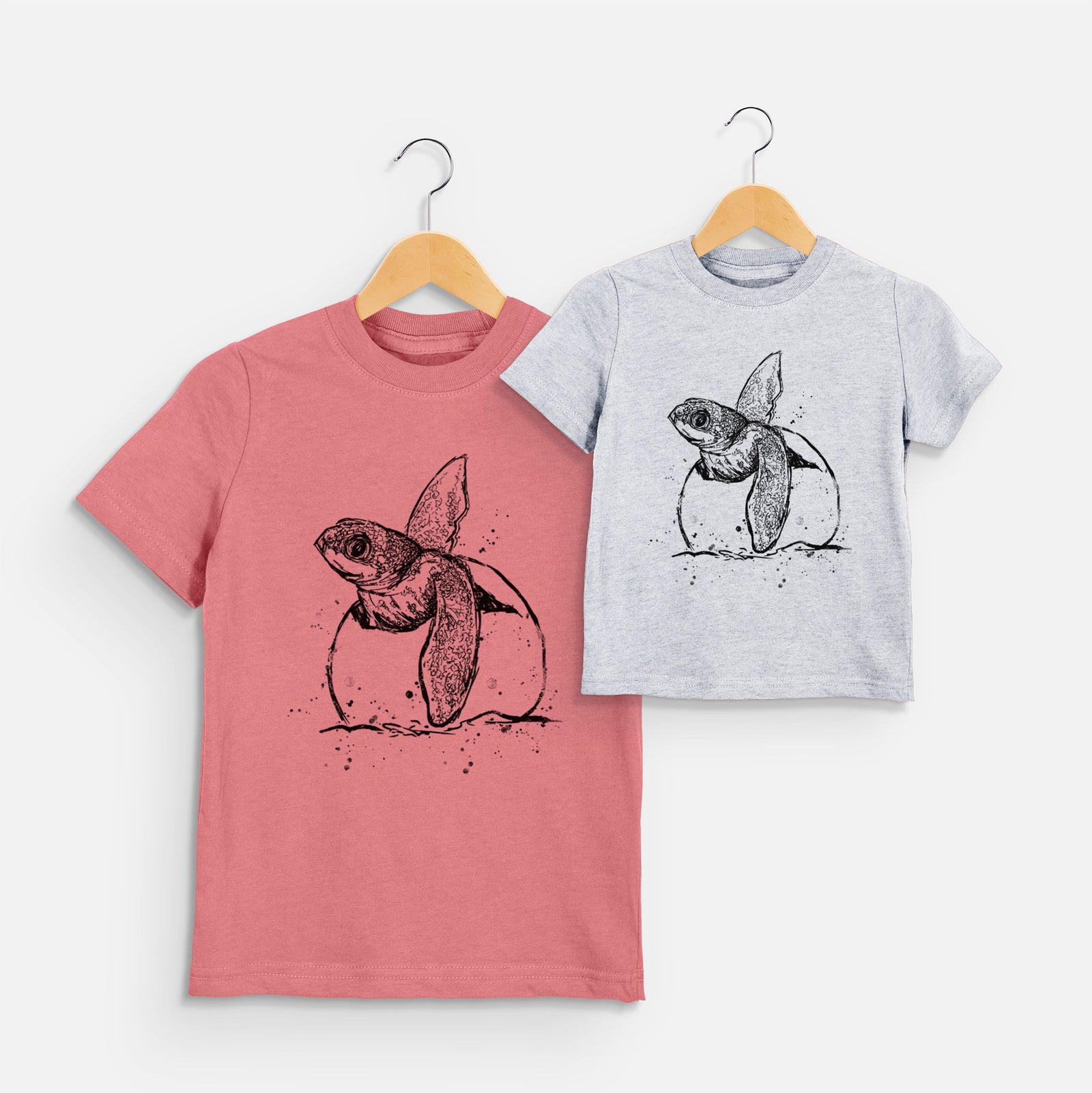 Two youth t-shirts in different colors on hangers, printed with a black ink illustration of a leatherback sea turtle hatchling emerging from its egg.