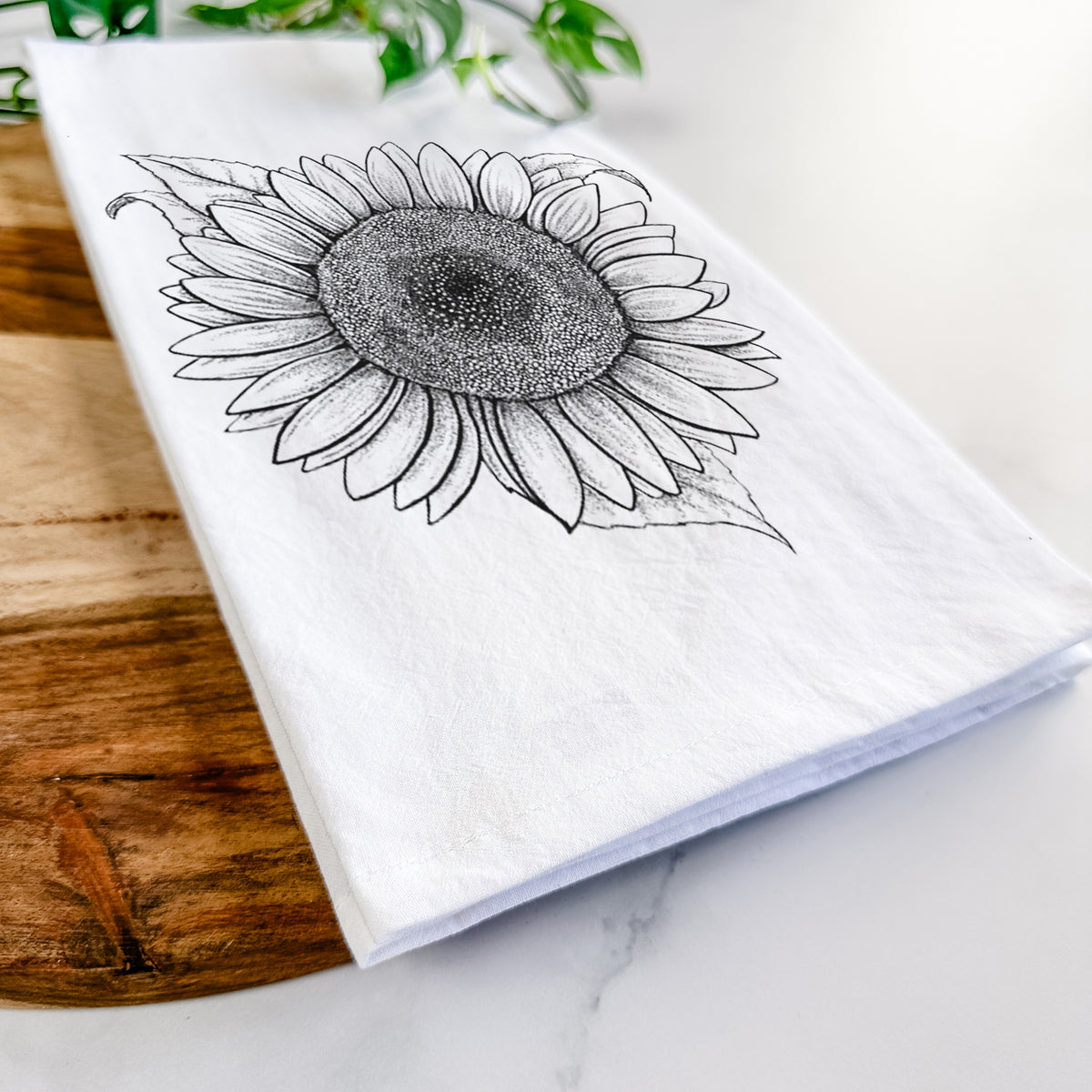 Close-up of a folded white tea towel on a wooden board, showing a detailed black ink line drawing of a Lemon Queen sunflower in bloom.