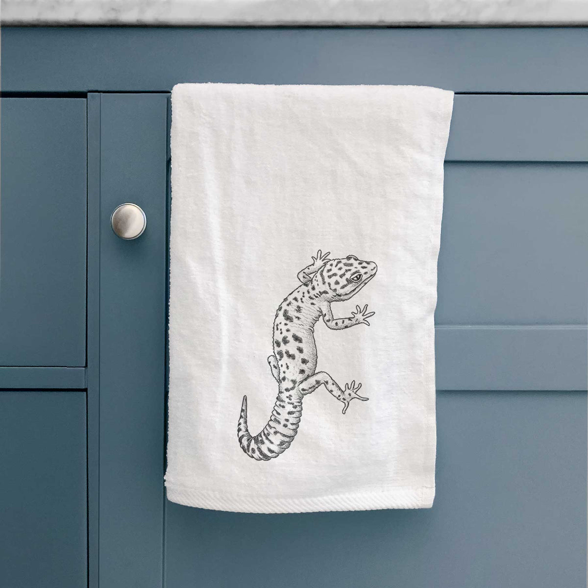 White hand towel printed with a detailed black ink illustration of a spotted leopard gecko climbing, hanging on a cabinet door.