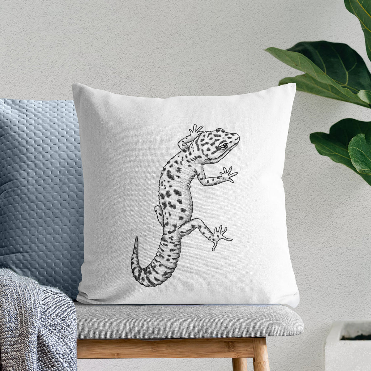 White pillow cover printed with a detailed black ink illustration of a spotted leopard gecko climbing, on a bench beside plants.