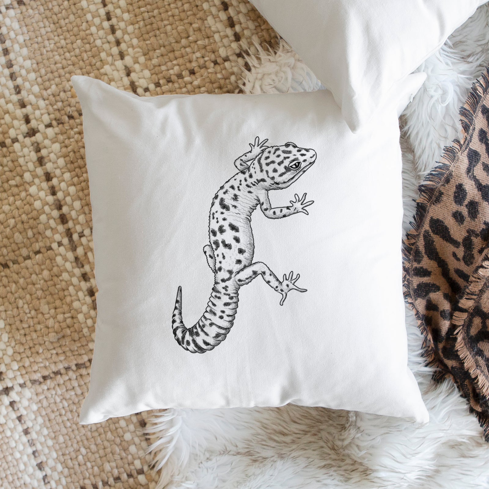 White pillow cover printed with a detailed black ink illustration of a spotted leopard gecko climbing, on a bench beside plants.