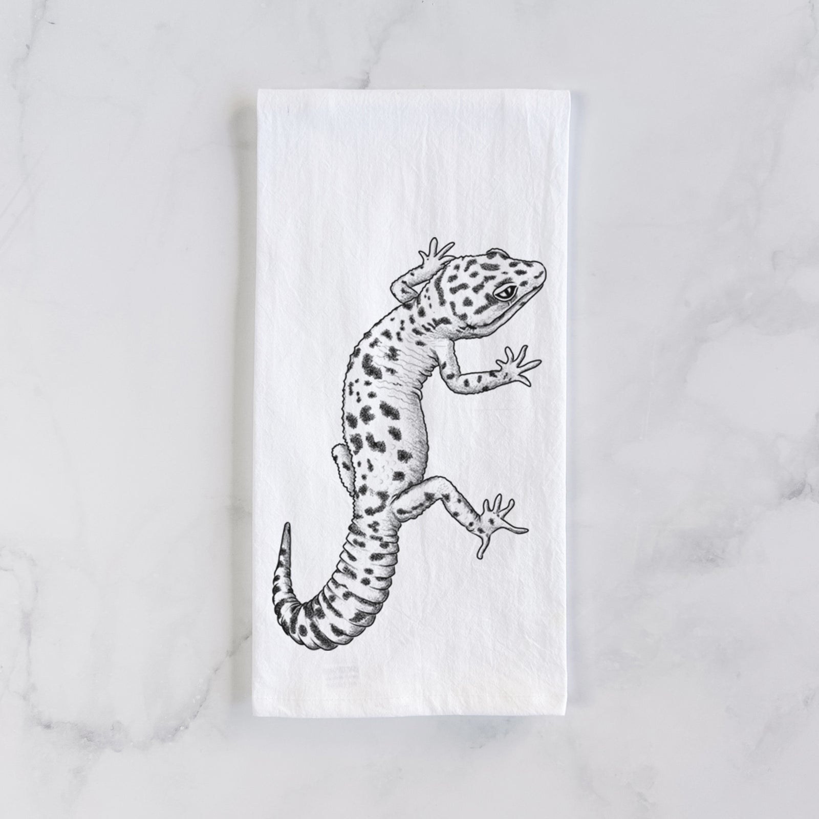 White cotton tea towel printed with a detailed black ink illustration of a spotted leopard gecko climbing, on a marble counter beside a round wooden cutting board and greenery.