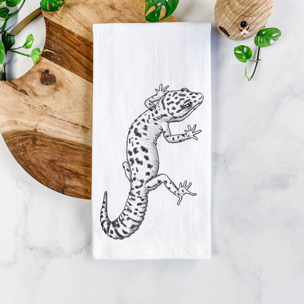 White cotton tea towel printed with a detailed black ink illustration of a spotted leopard gecko climbing, on a marble counter beside a round wooden cutting board and greenery.