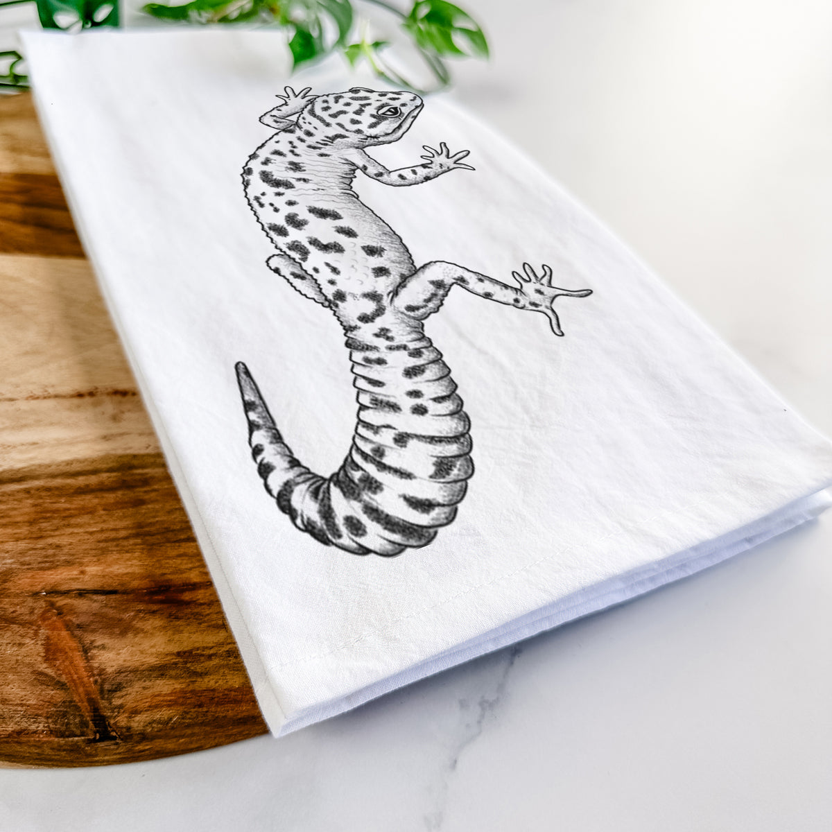 Close-up of a folded white tea towel on a wooden board, showing a detailed black ink illustration of a spotted leopard gecko climbing.