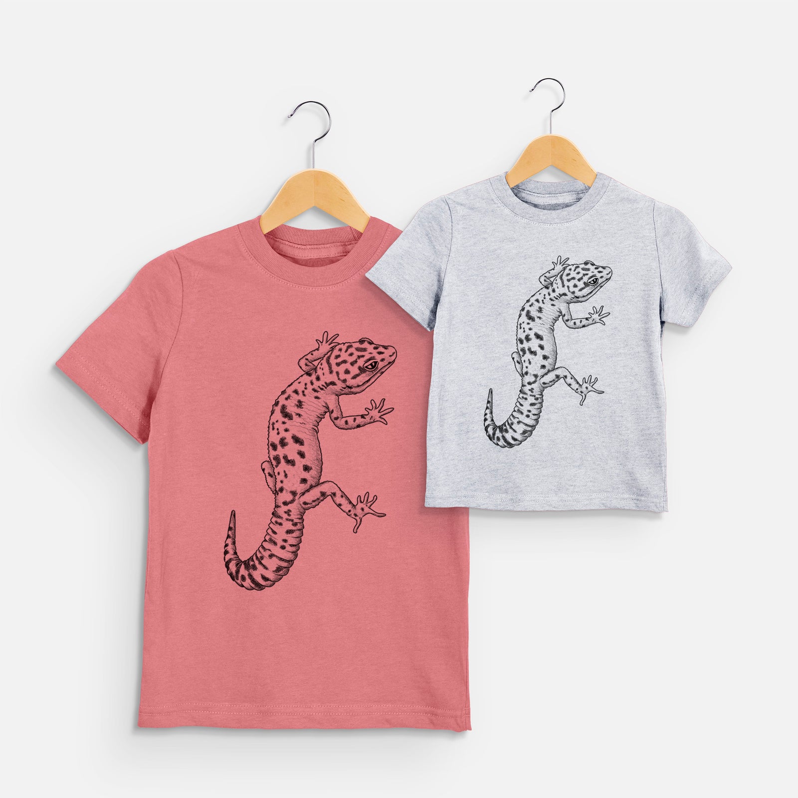 Two youth t-shirts in different colors on hangers, printed with a detailed black ink illustration of a spotted leopard gecko climbing.