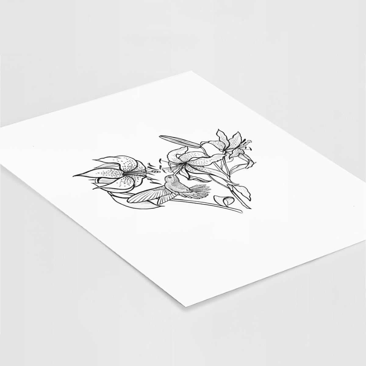 Art print of a black ink line drawing of lilies and a hummingbird arranged in a heart shape, lying at an angle on a white surface.