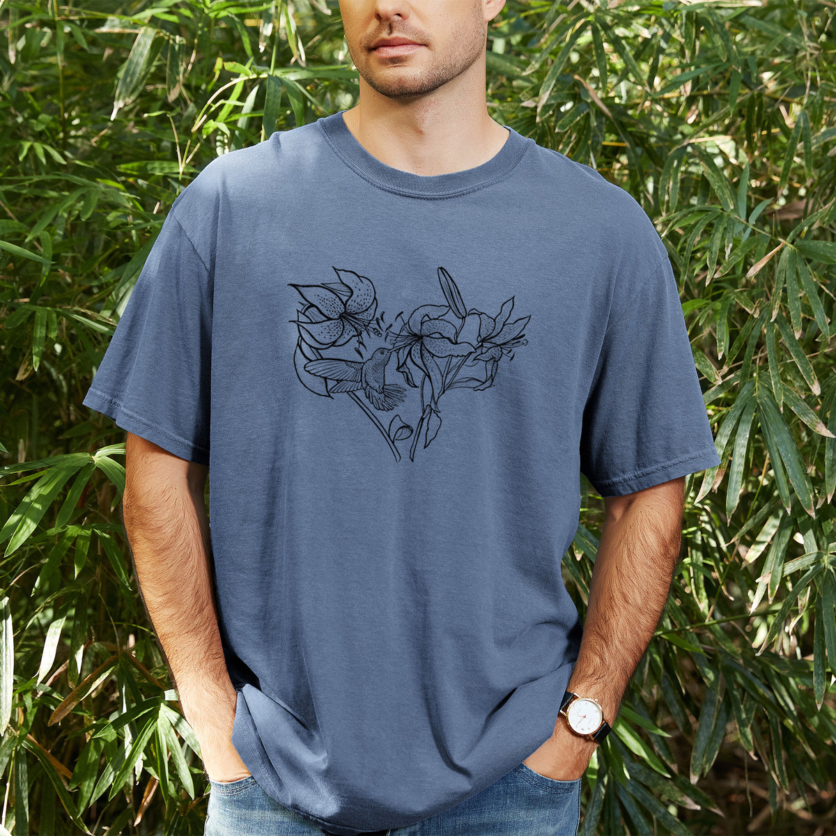 Man wearing a Comfort Colors t-shirt printed with a black ink line drawing of lilies and a hummingbird arranged in a heart shape, outdoors.