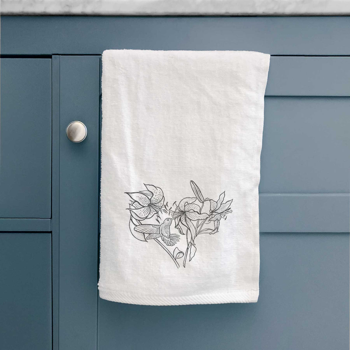 White hand towel printed with a black ink line drawing of lilies and a hummingbird arranged in a heart shape, hanging on a cabinet door.