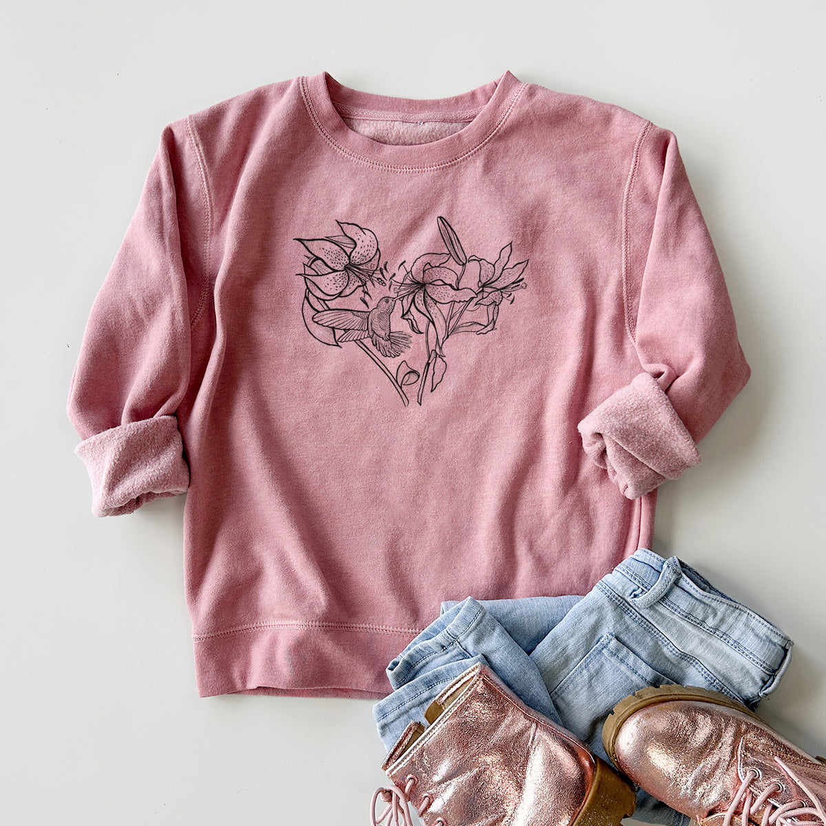 Youth crewneck sweatshirt printed with a black ink line drawing of lilies and a hummingbird arranged in a heart shape, styled flat with jeans and boots.