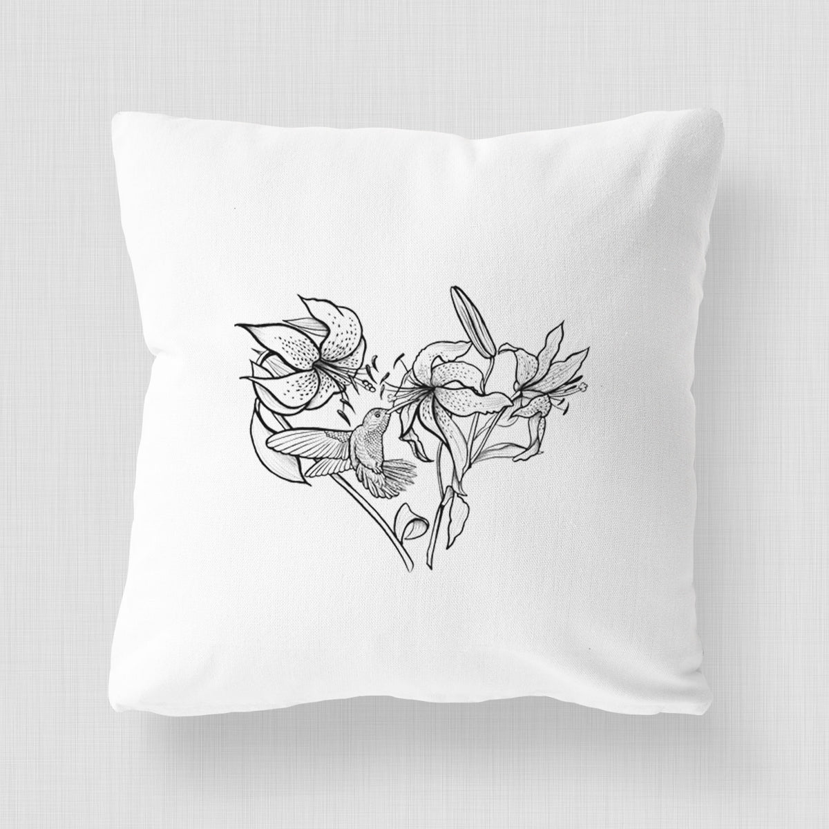 White pillow cover printed with a black ink line drawing of lilies and a hummingbird arranged in a heart shape.