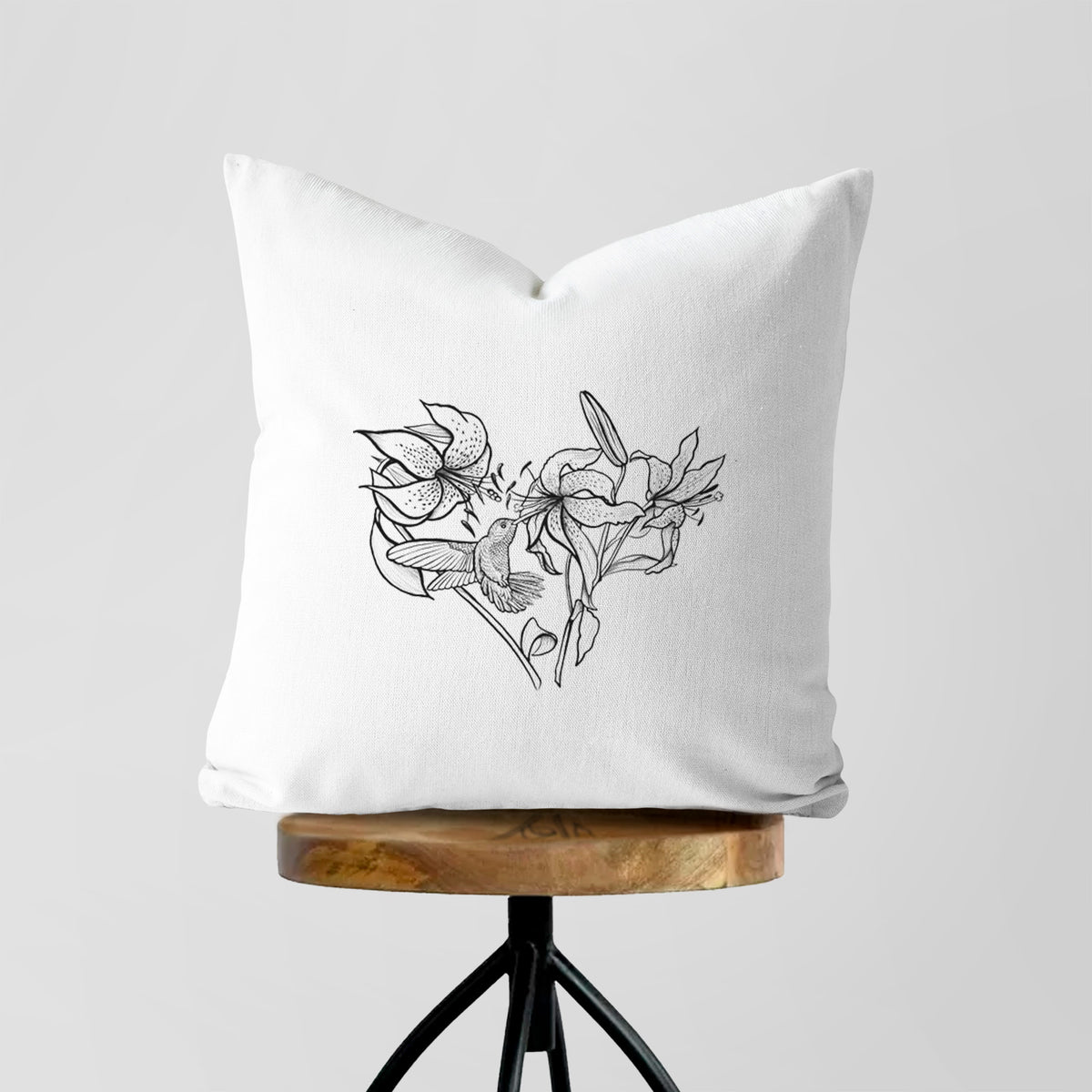 White pillow cover printed with a black ink line drawing of lilies and a hummingbird arranged in a heart shape, on a wooden stool.