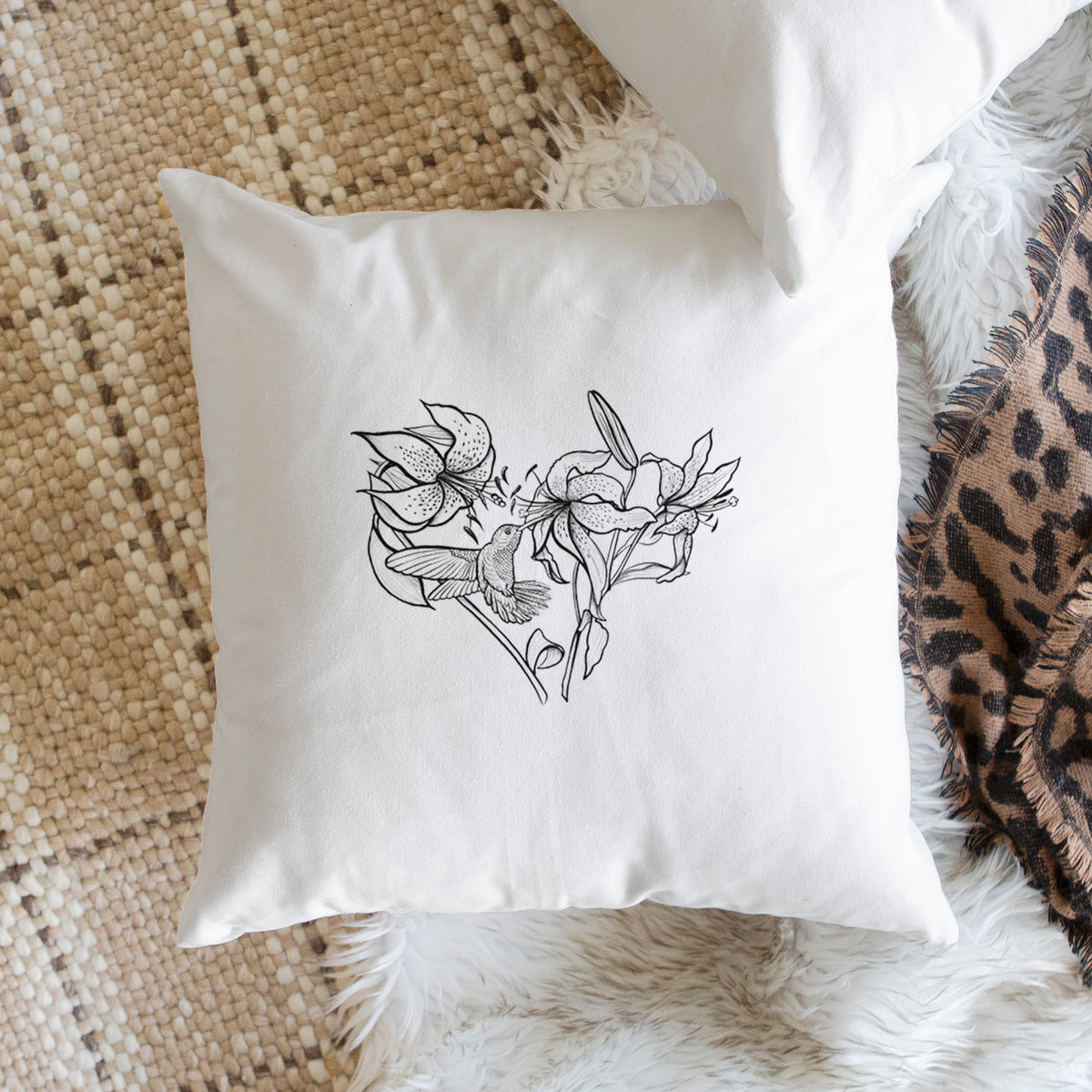 White pillow cover printed with a black ink line drawing of lilies and a hummingbird arranged in a heart shape, on a woven chair.