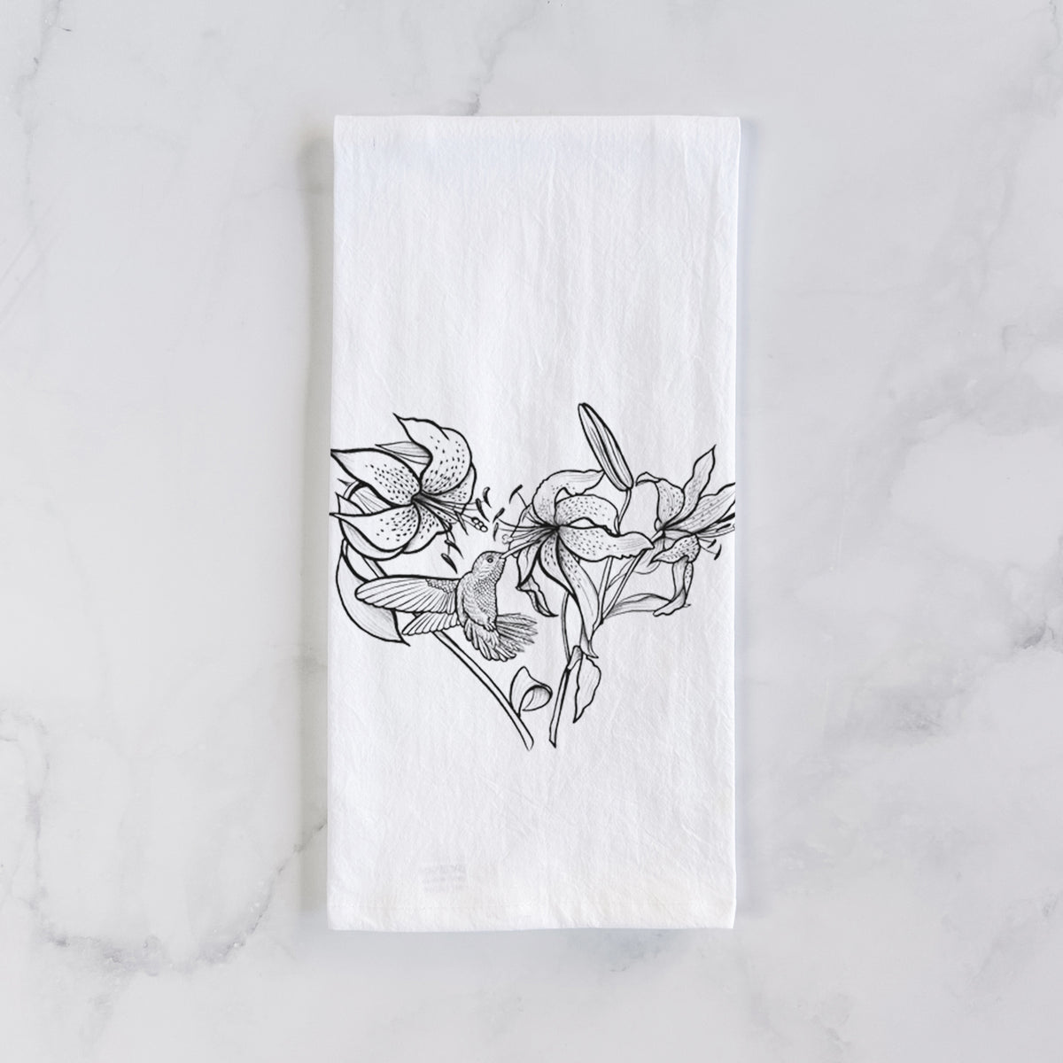 White cotton tea towel laid flat on marble, printed with a black ink line drawing of lilies and a hummingbird arranged in a heart shape.