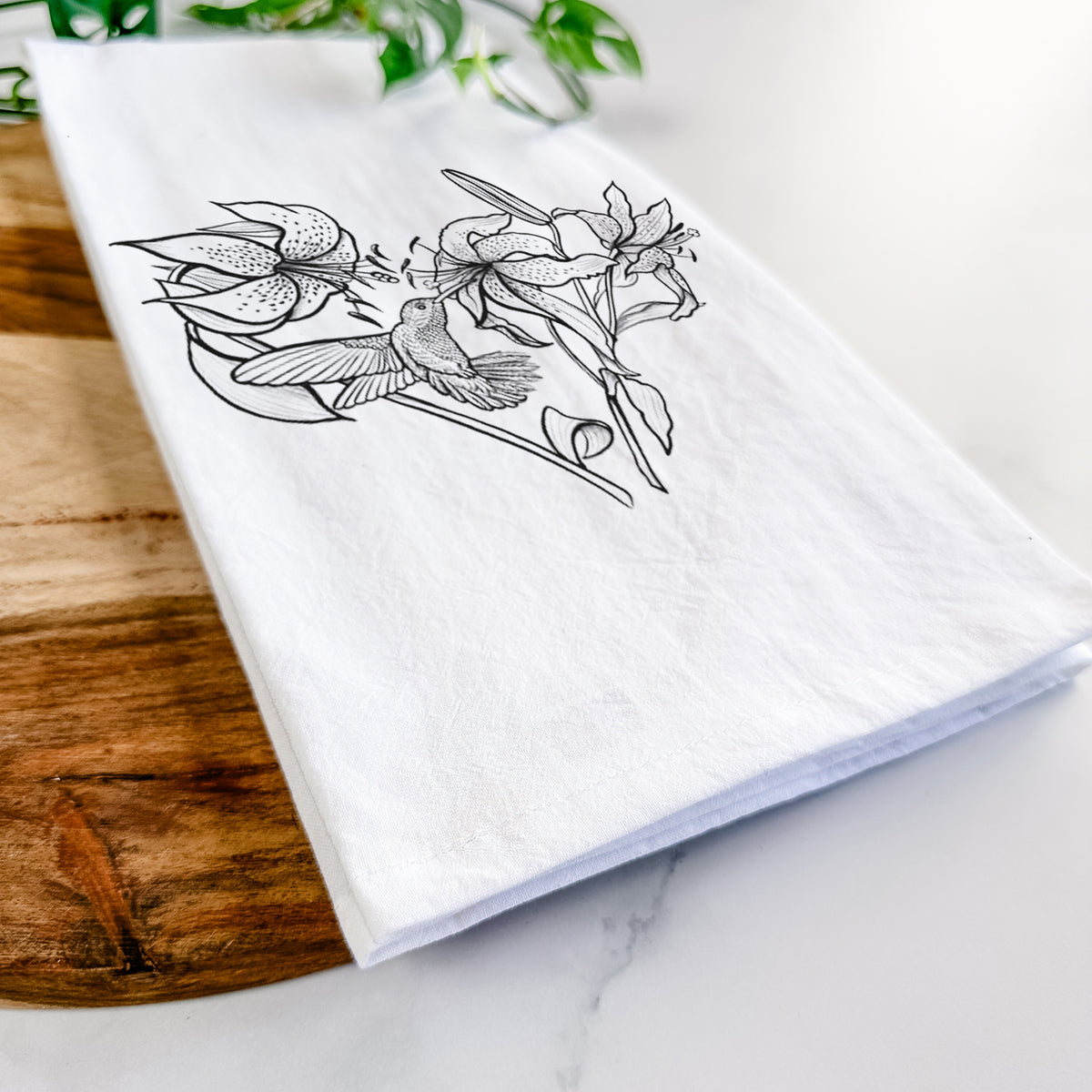 Close-up of a folded white tea towel on a wooden board, showing a black ink line drawing of lilies and a hummingbird arranged in a heart shape.