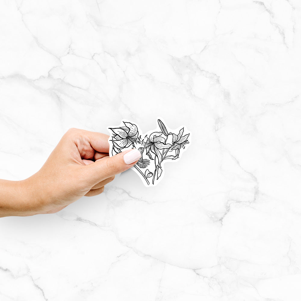 Hand holding a 4 inch vinyl decal of a black ink line drawing of lilies and a hummingbird arranged in a heart shape.