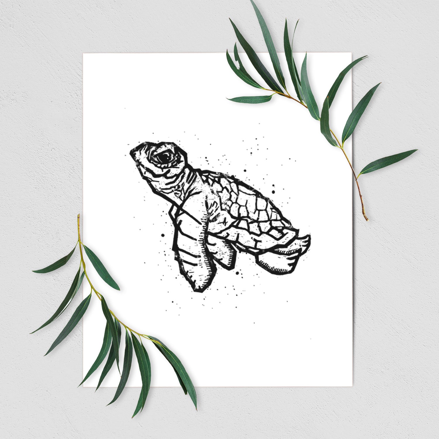 Art print of a black linocut-style illustration of a baby sea turtle with a patterned shell and ink splatters, shown with eucalyptus leaves.