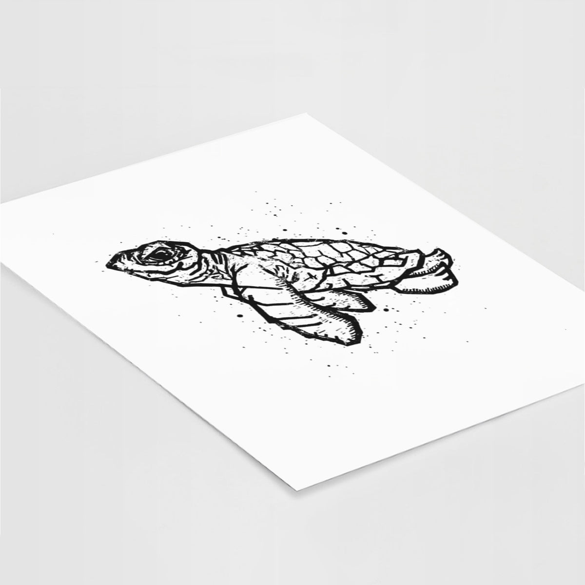 Art print of a black linocut-style illustration of a baby sea turtle with a patterned shell and ink splatters, lying at an angle on a white surface.