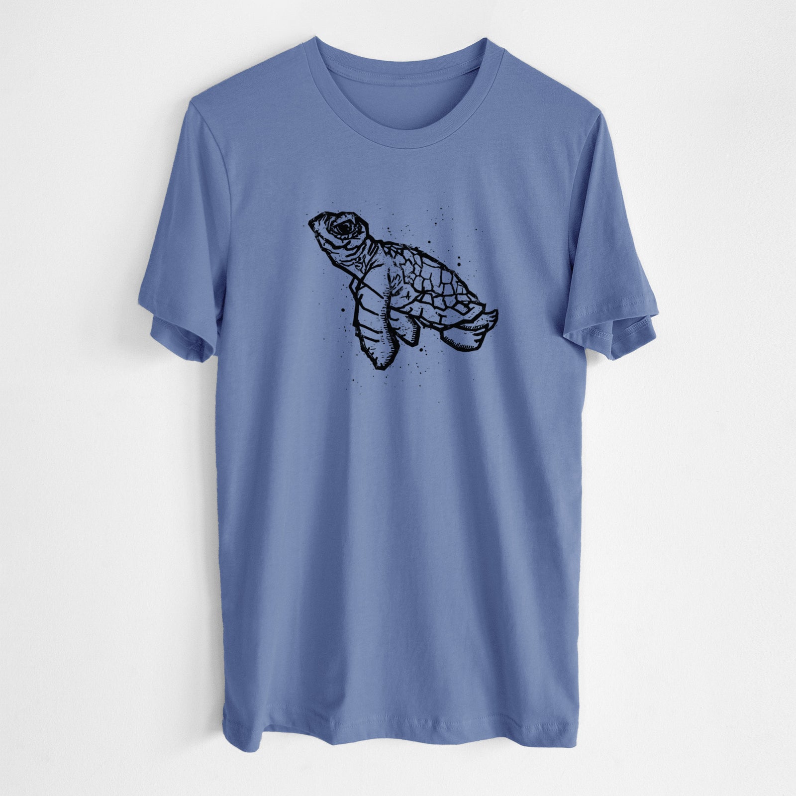 Spring green lightweight cotton t-shirt laid flat with the sleeves rolled, printed with a black linocut-style illustration of a baby sea turtle with a patterned shell and ink splatters.