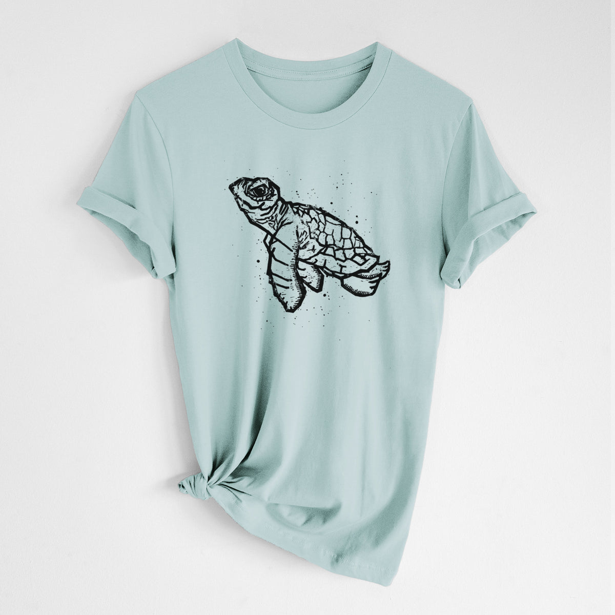 Dusty blue lightweight cotton t-shirt laid flat with the sleeves rolled, printed with a black linocut-style illustration of a baby sea turtle with a patterned shell and ink splatters.