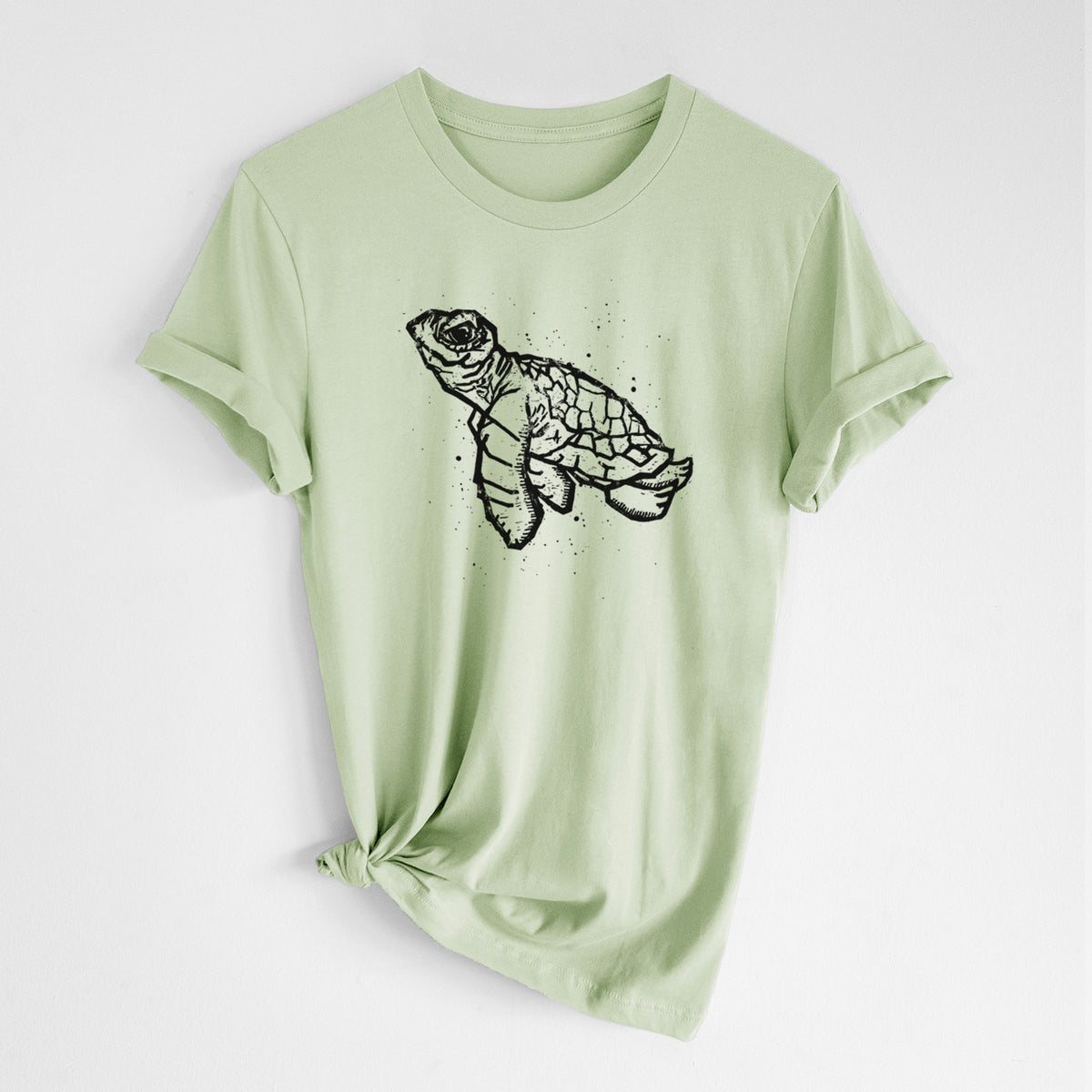 Spring green lightweight cotton t-shirt laid flat with the sleeves rolled, printed with a black linocut-style illustration of a baby sea turtle with a patterned shell and ink splatters.