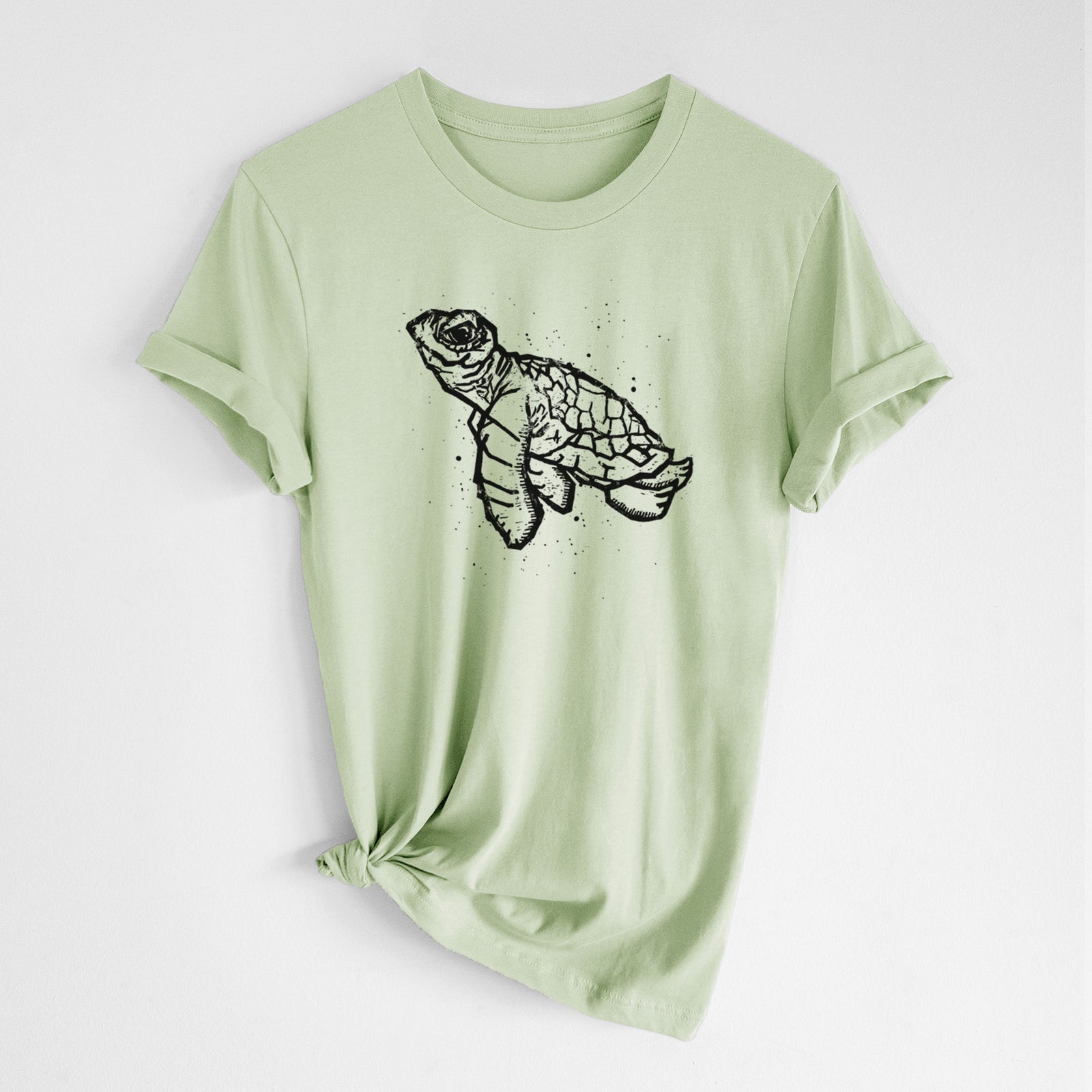 Spring green lightweight cotton t-shirt laid flat with the sleeves rolled, printed with a black linocut-style illustration of a baby sea turtle with a patterned shell and ink splatters.