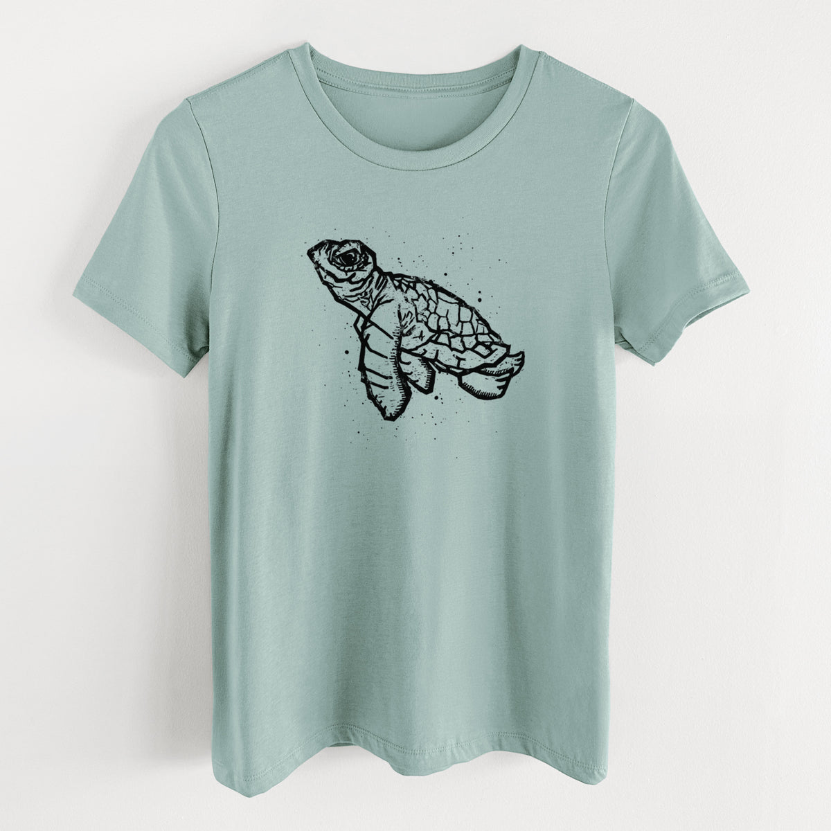 Dusty blue women's relaxed fit cotton t-shirt laid flat, printed with a black linocut-style illustration of a baby sea turtle with a patterned shell and ink splatters.