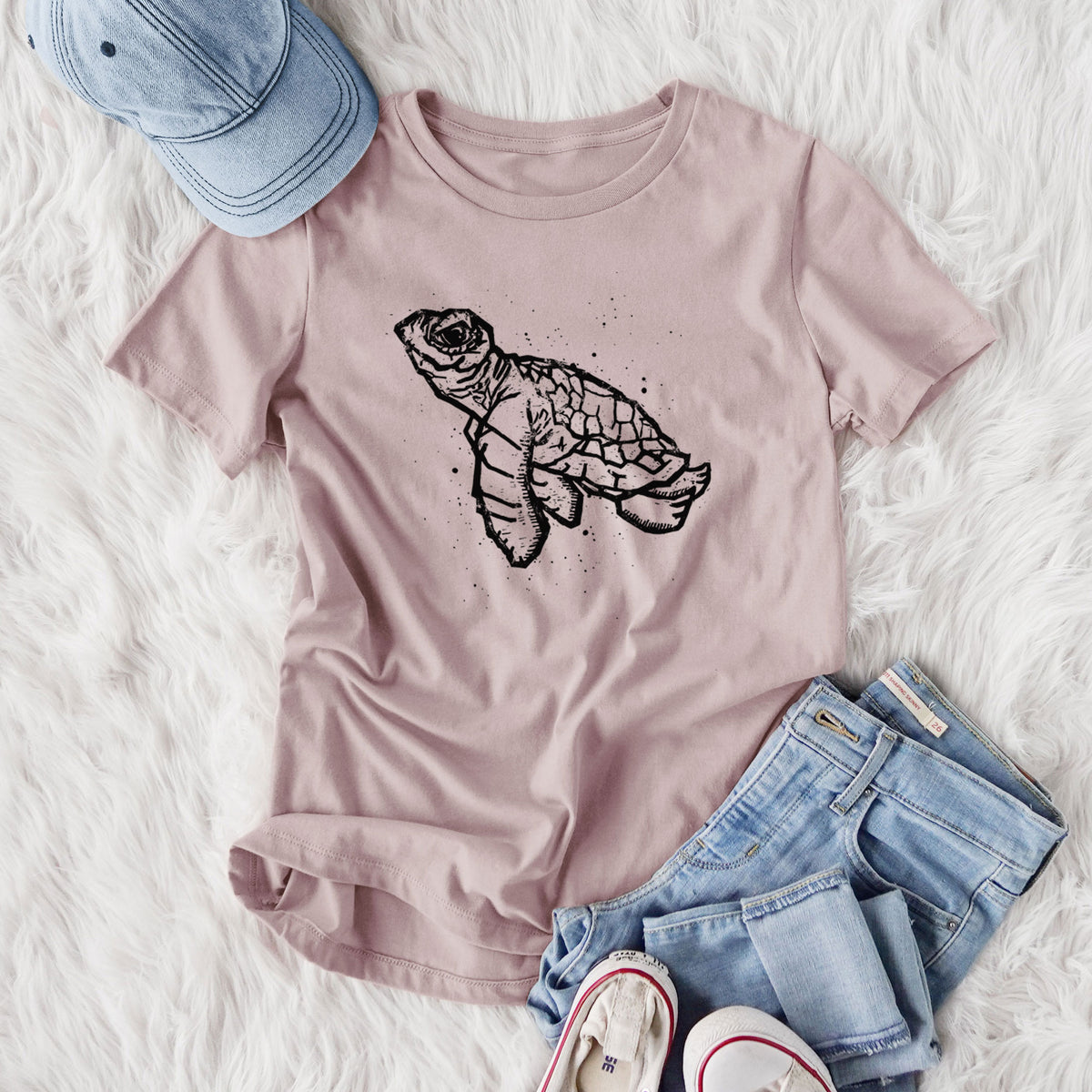 Women's relaxed fit cotton t-shirt printed with a black linocut-style illustration of a baby sea turtle with a patterned shell and ink splatters, styled flat on a white faux fur rug with jeans.
