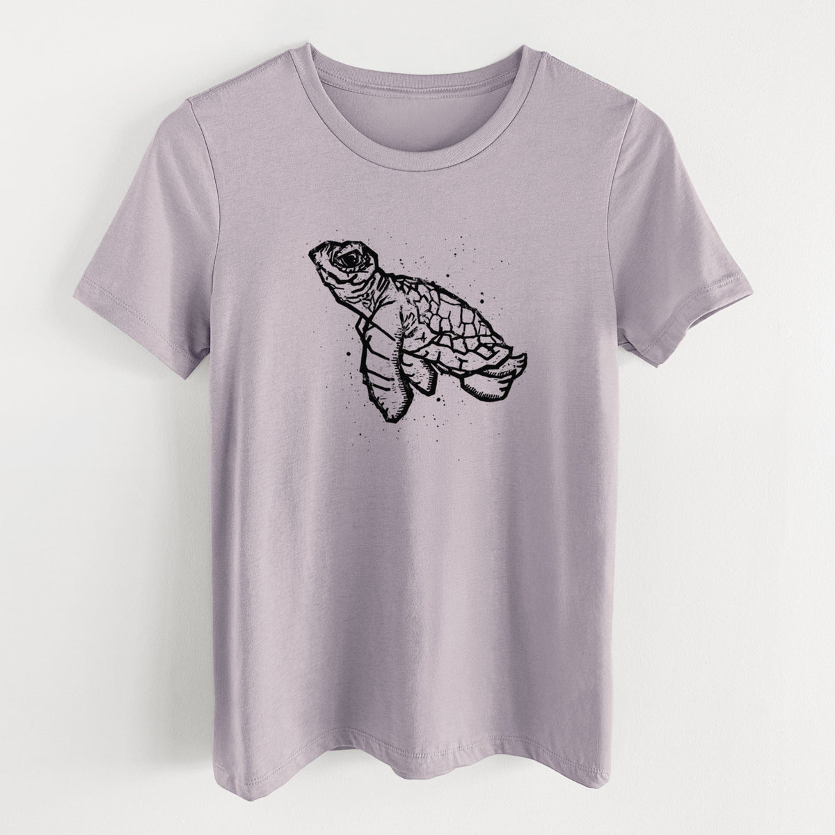 Light violet women's relaxed fit cotton t-shirt laid flat, printed with a black linocut-style illustration of a baby sea turtle with a patterned shell and ink splatters.
