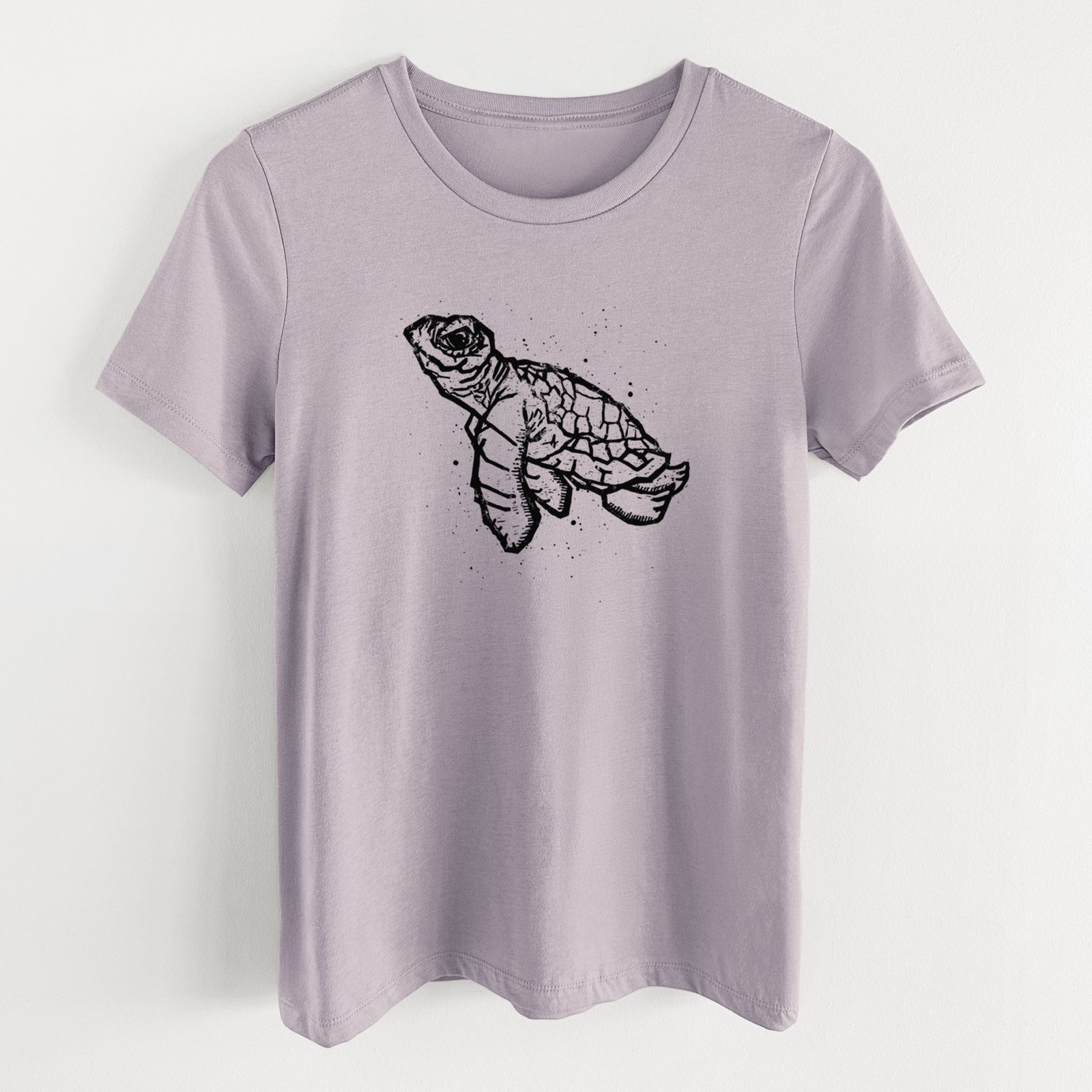 Light violet women's relaxed fit cotton t-shirt laid flat, printed with a black linocut-style illustration of a baby sea turtle with a patterned shell and ink splatters.