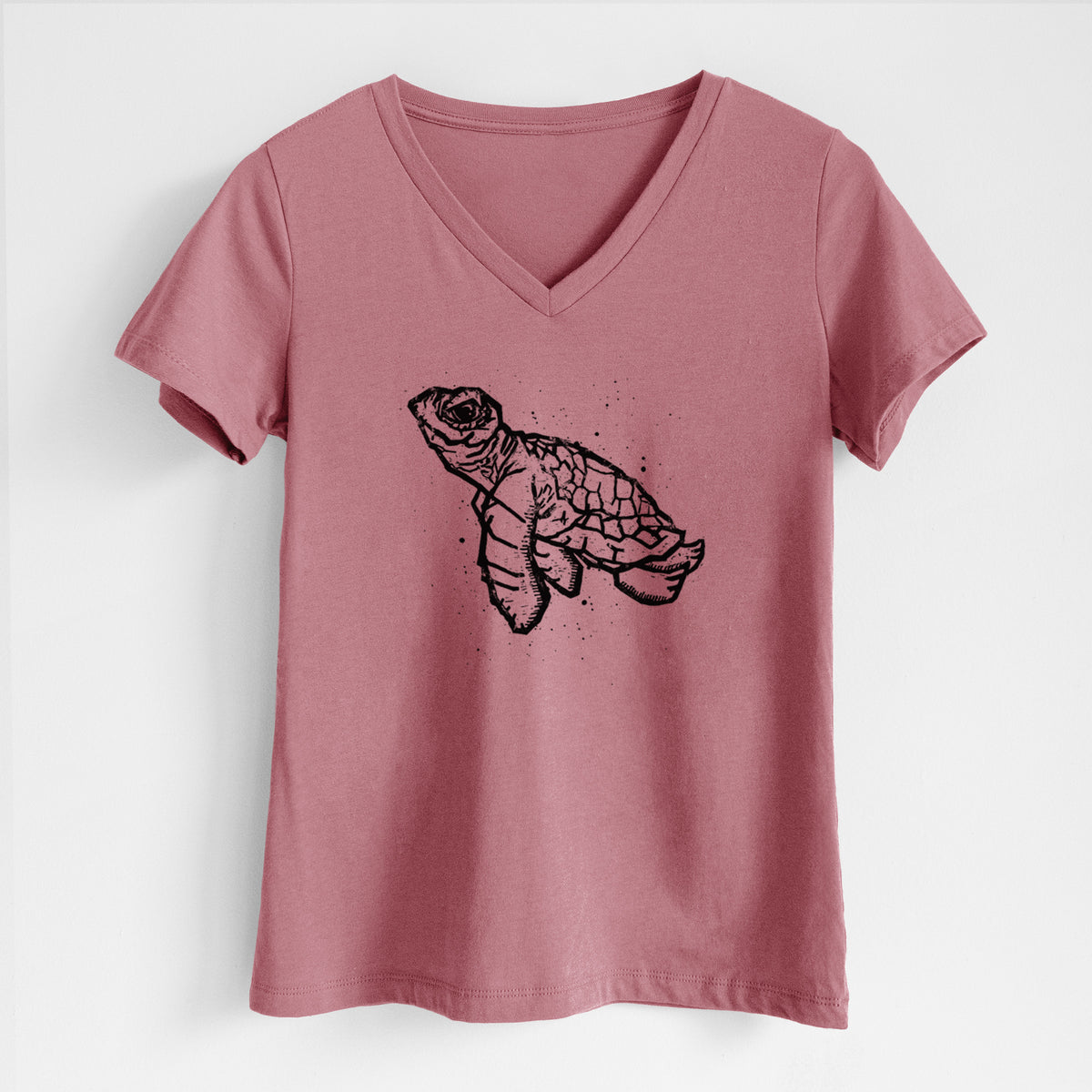 Mauve pink women's relaxed fit cotton V-neck t-shirt laid flat, printed with a black linocut-style illustration of a baby sea turtle with a patterned shell and ink splatters.