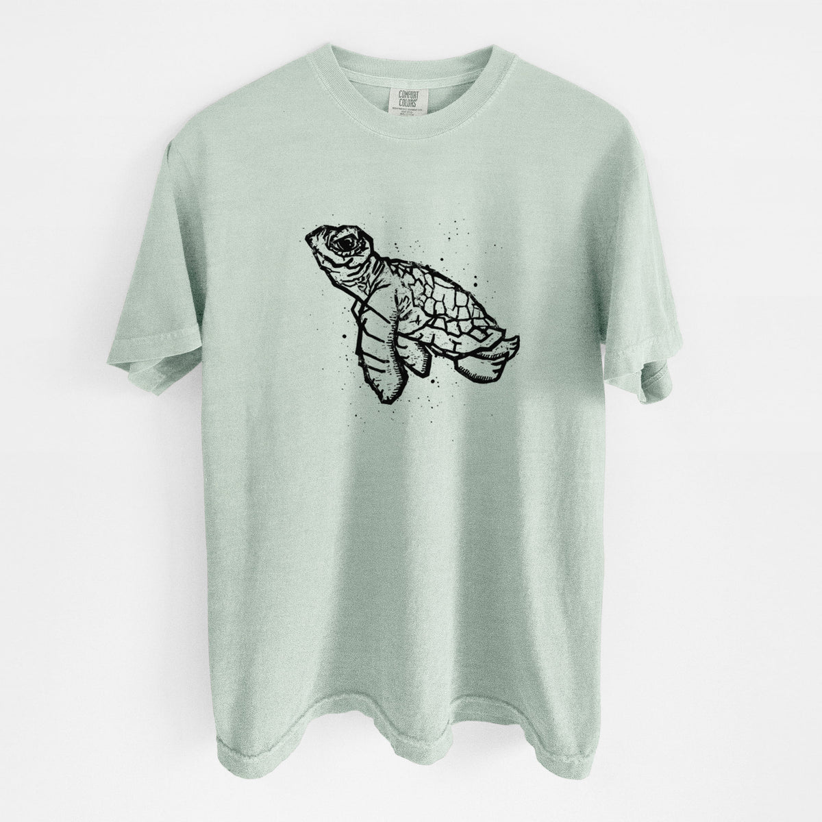 Bay green Comfort Colors t-shirt laid flat, printed with a black linocut-style illustration of a baby sea turtle with a patterned shell and ink splatters.