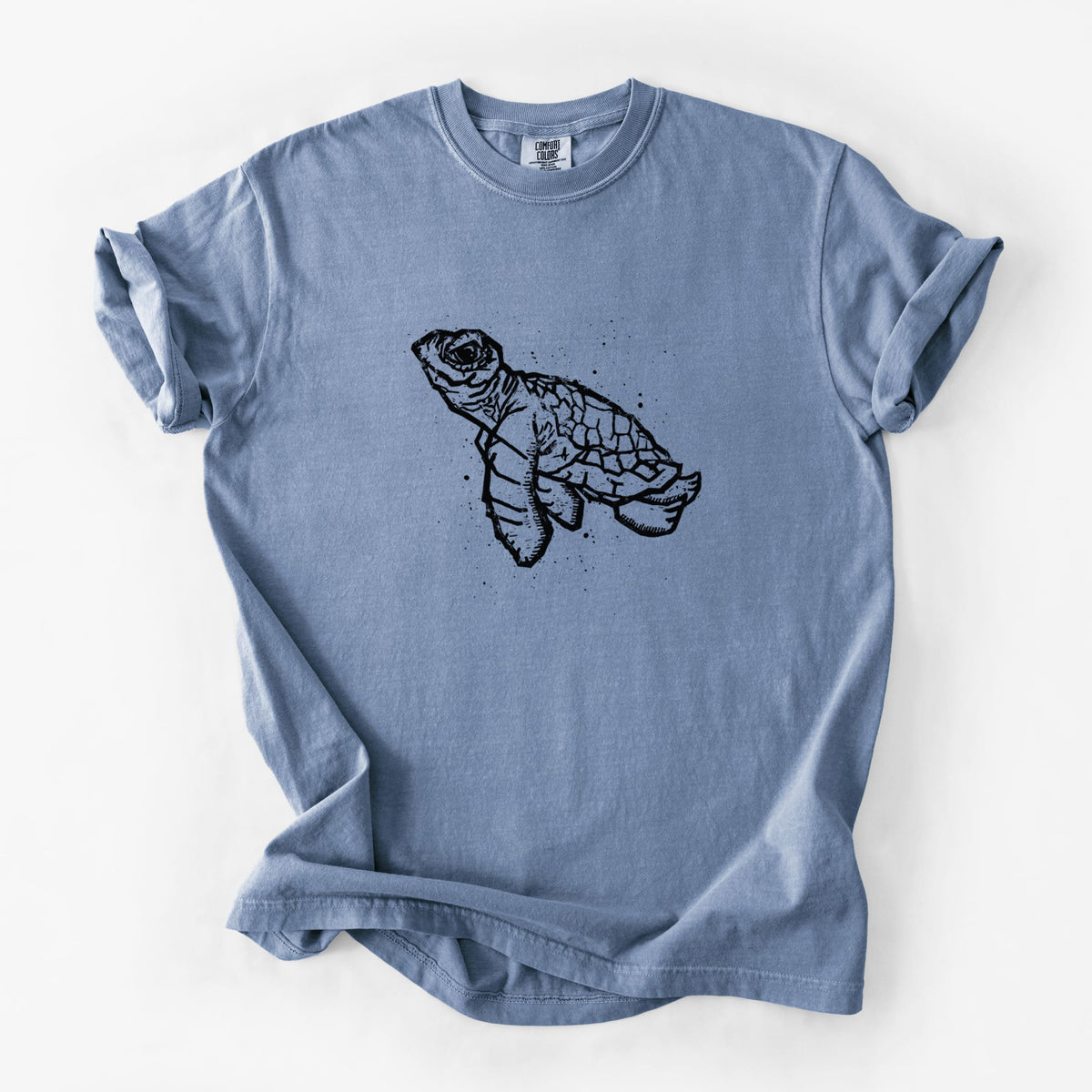 Blue jean Comfort Colors t-shirt laid flat, printed with a black linocut-style illustration of a baby sea turtle with a patterned shell and ink splatters.