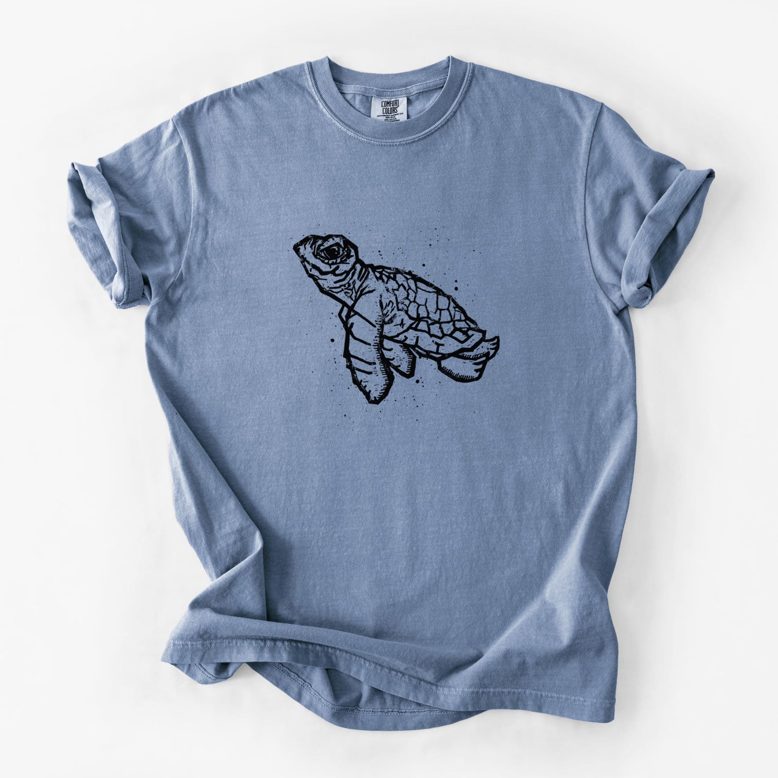 Blue jean Comfort Colors t-shirt laid flat, printed with a black linocut-style illustration of a baby sea turtle with a patterned shell and ink splatters.