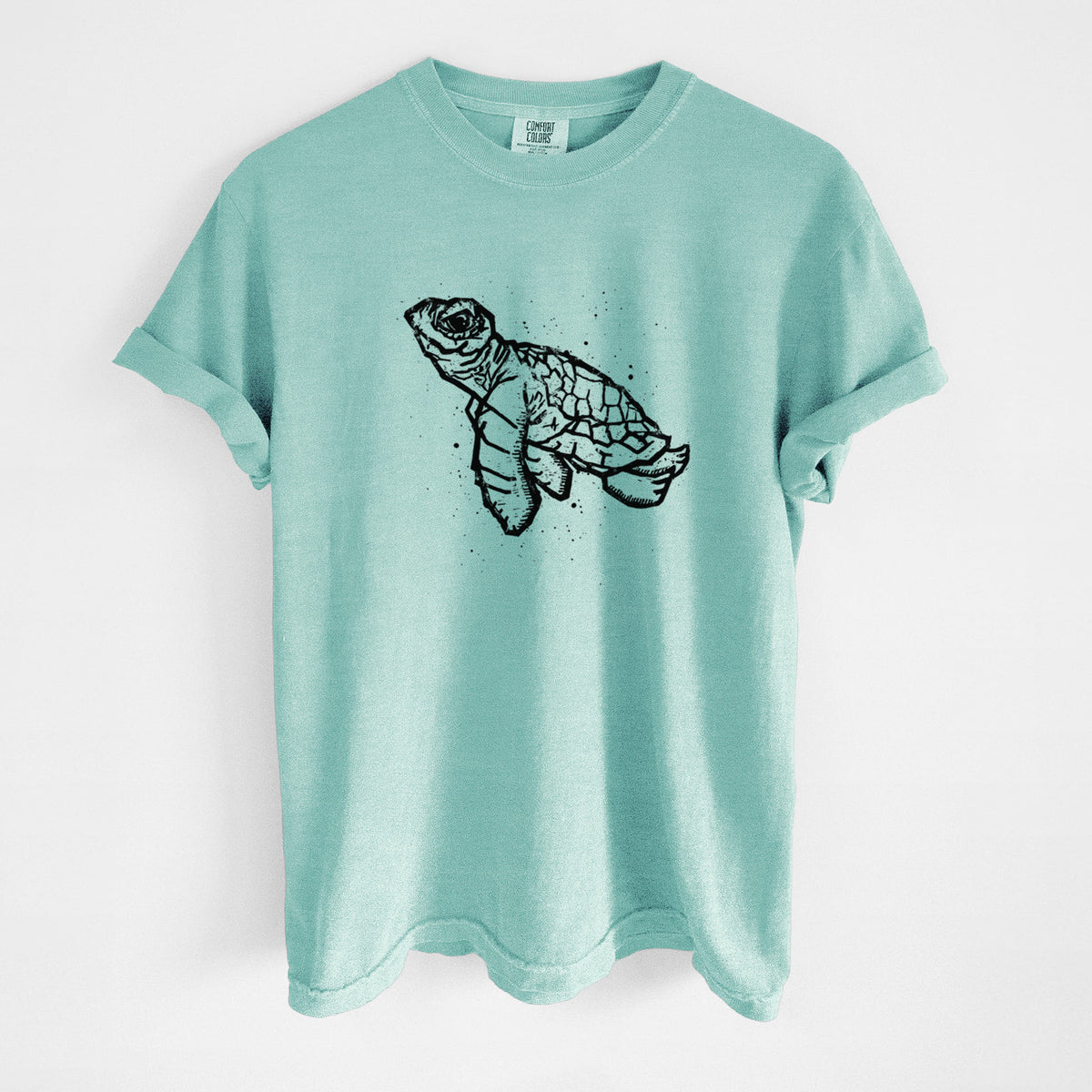 Chalky mint Comfort Colors t-shirt laid flat, printed with a black linocut-style illustration of a baby sea turtle with a patterned shell and ink splatters.