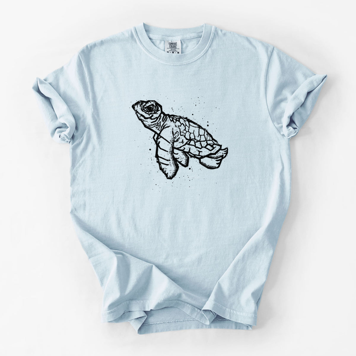 Chambray blue Comfort Colors t-shirt laid flat, printed with a black linocut-style illustration of a baby sea turtle with a patterned shell and ink splatters.