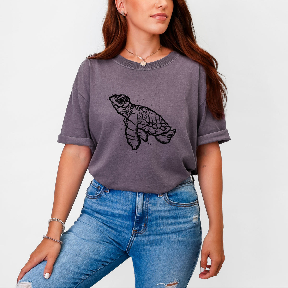 Woman wearing a Comfort Colors t-shirt printed with a black linocut-style illustration of a baby sea turtle with a patterned shell and ink splatters.
