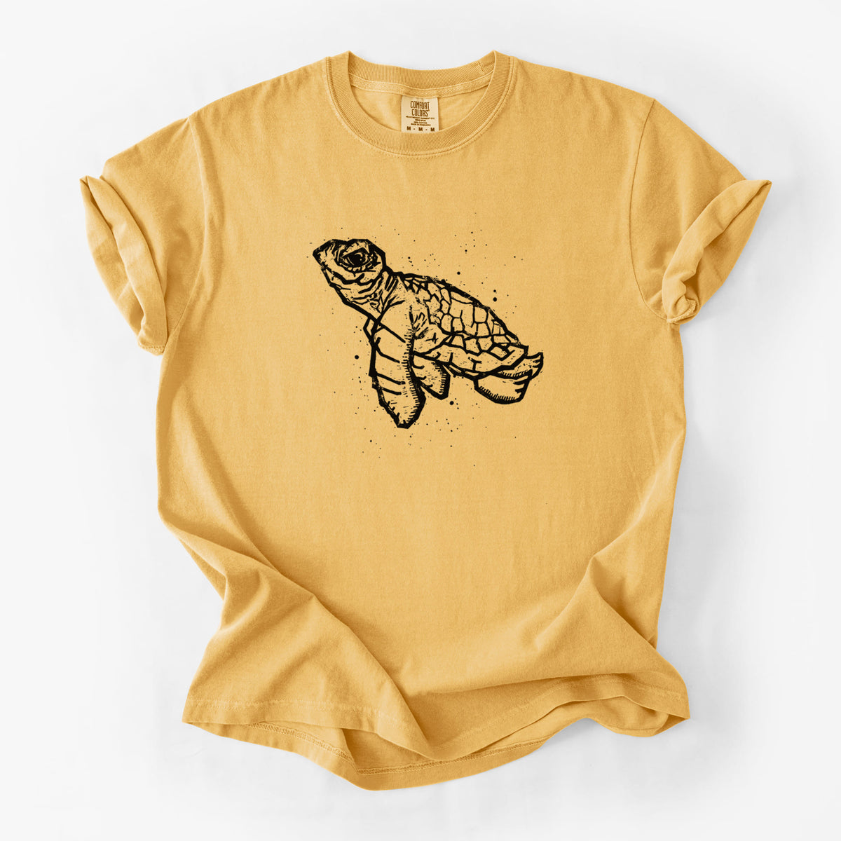 Golden wheat yellow Comfort Colors t-shirt laid flat, printed with a black linocut-style illustration of a baby sea turtle with a patterned shell and ink splatters.