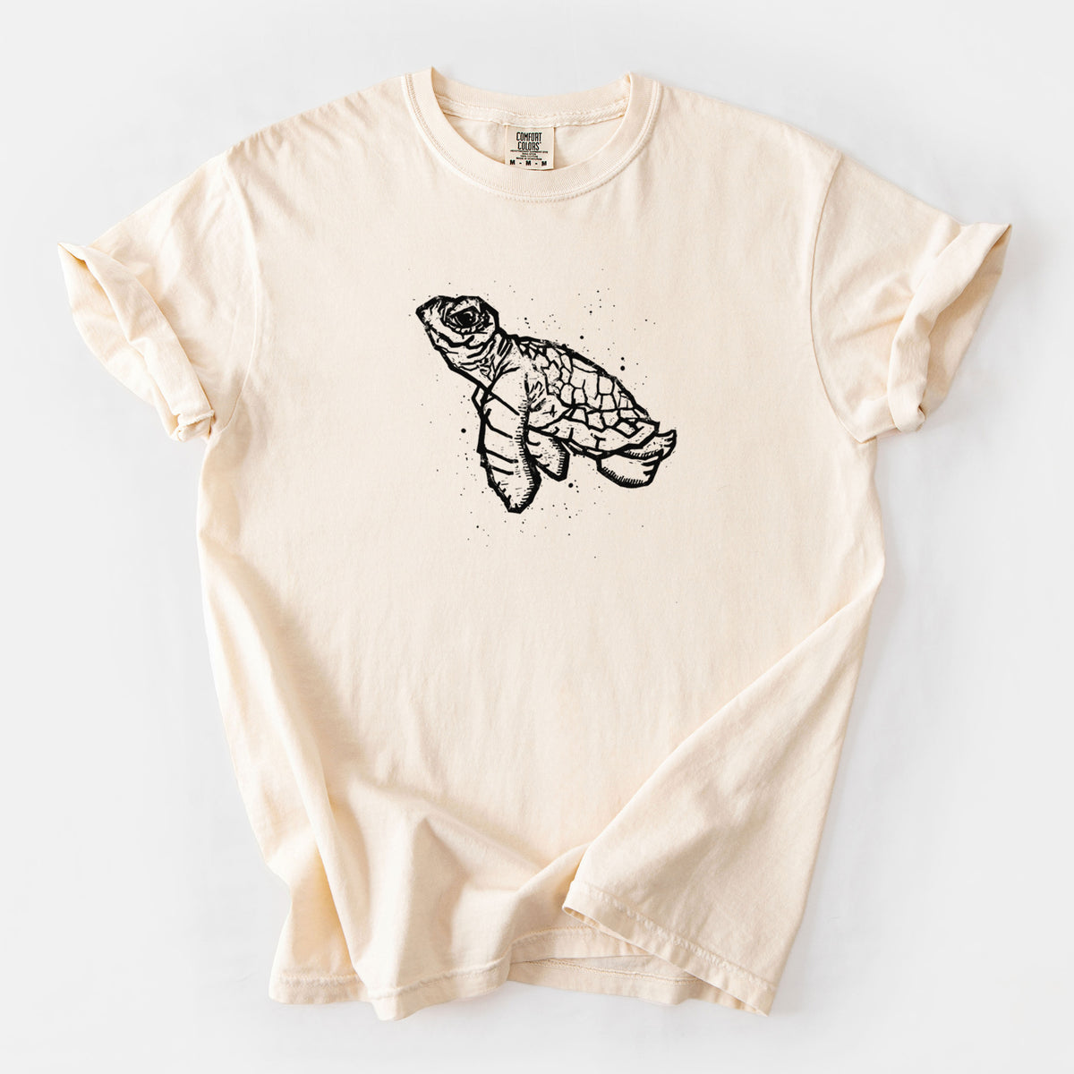 Ivory Comfort Colors t-shirt laid flat, printed with a black linocut-style illustration of a baby sea turtle with a patterned shell and ink splatters.