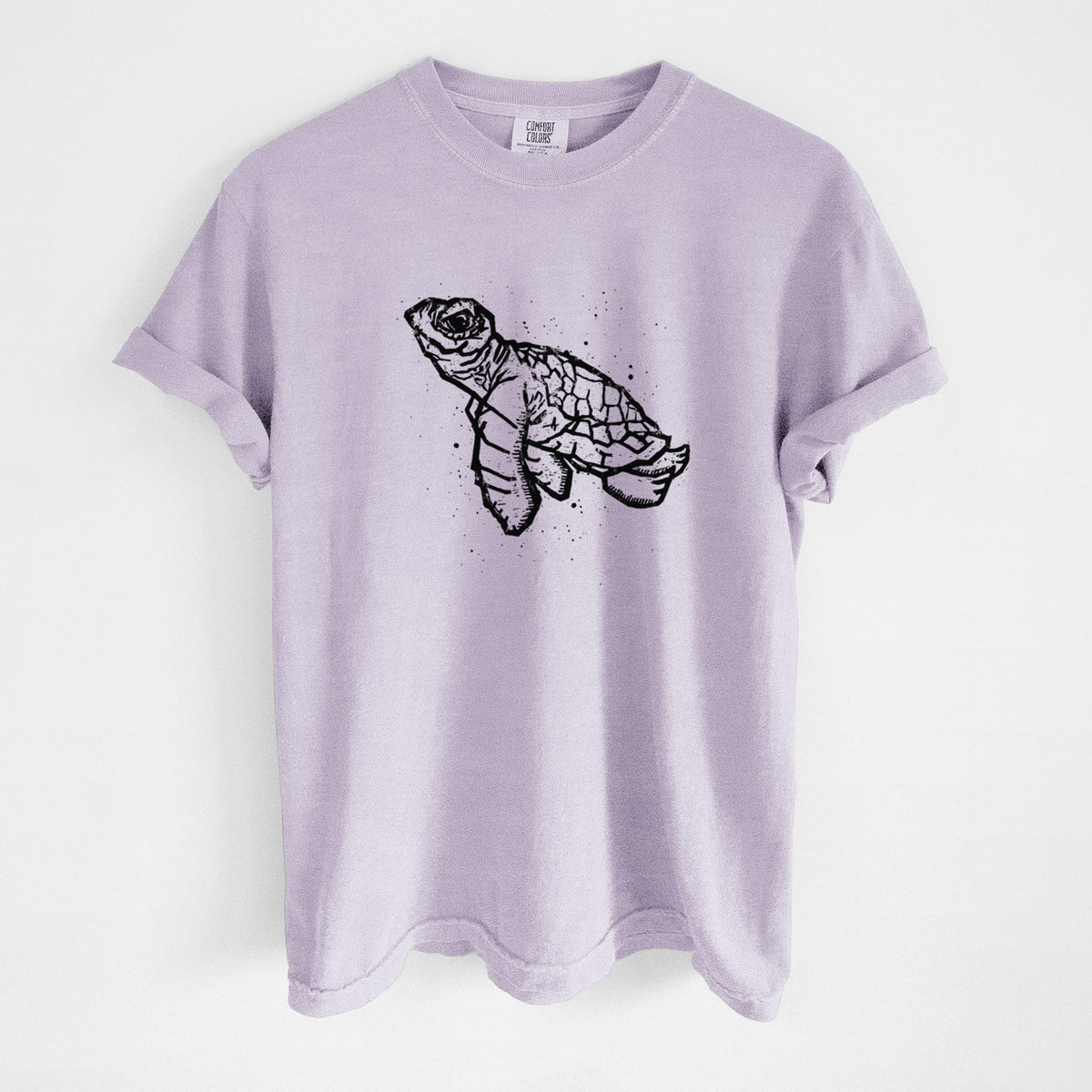 Orchid purple Comfort Colors t-shirt laid flat, printed with a black linocut-style illustration of a baby sea turtle with a patterned shell and ink splatters.
