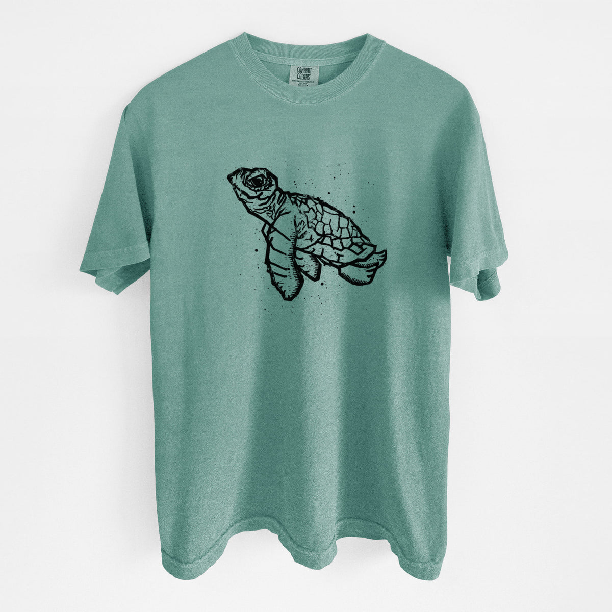Reef green Comfort Colors t-shirt laid flat, printed with a black linocut-style illustration of a baby sea turtle with a patterned shell and ink splatters.