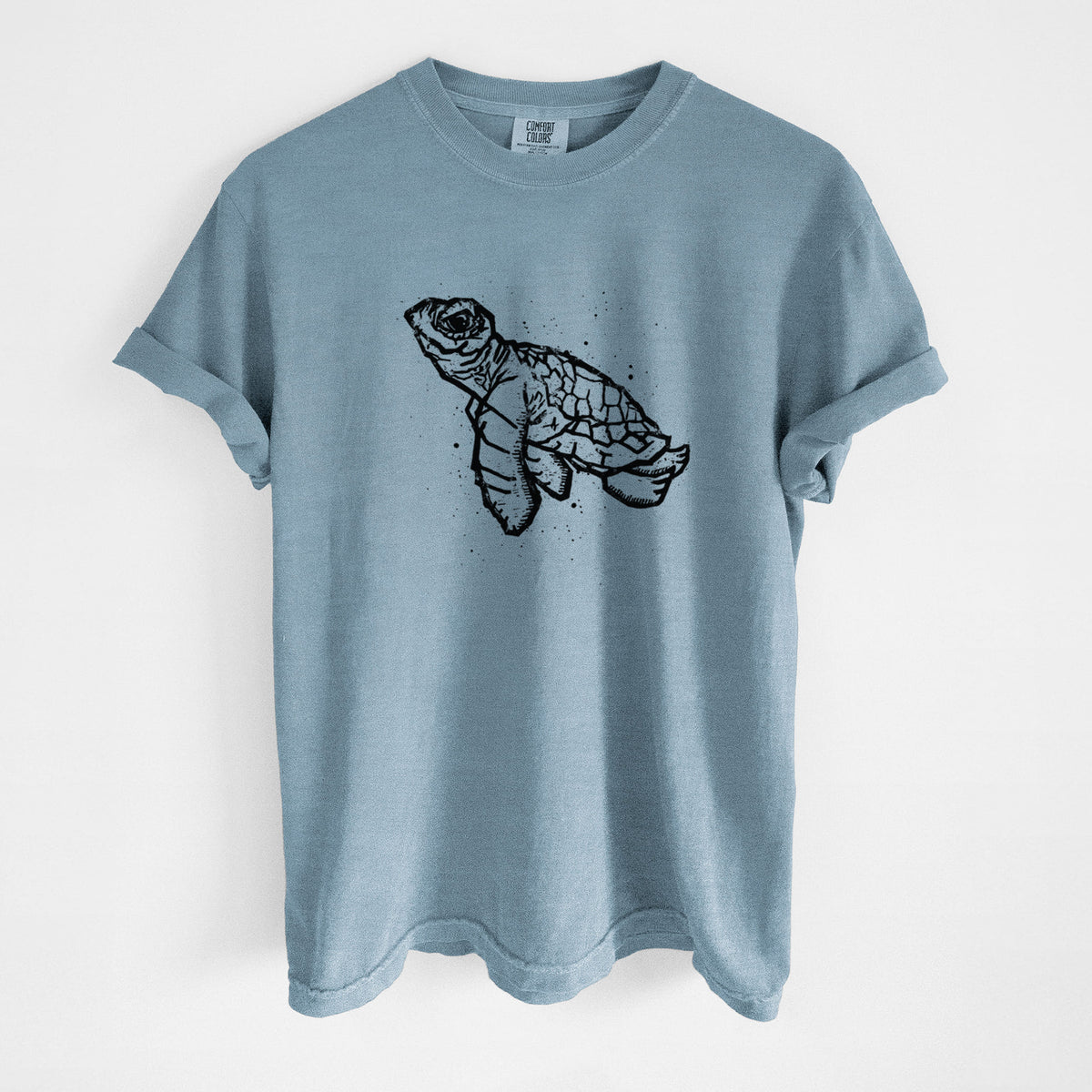 Steel blue Comfort Colors t-shirt laid flat, printed with a black linocut-style illustration of a baby sea turtle with a patterned shell and ink splatters.