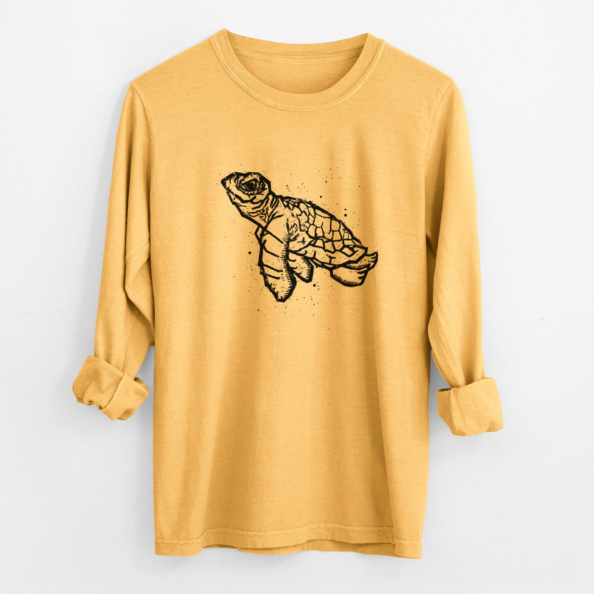 Golden wheat yellow Comfort Colors long sleeve shirt laid flat with the cuffs rolled, printed with a black linocut-style illustration of a baby sea turtle with a patterned shell and ink splatters.