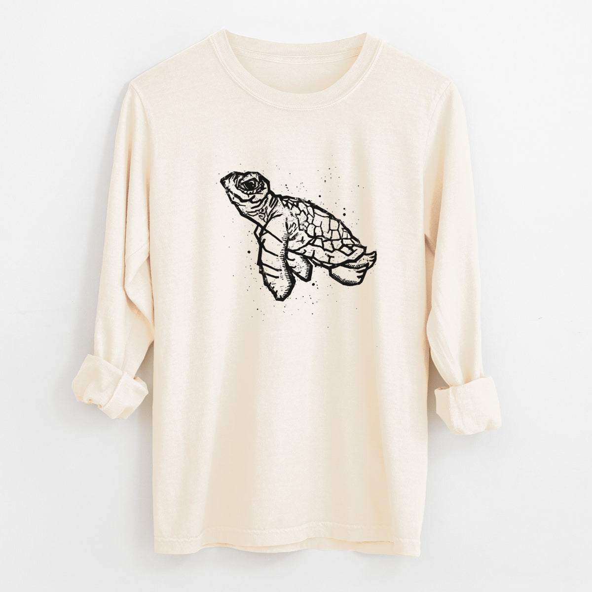 Ivory Comfort Colors long sleeve shirt laid flat with the cuffs rolled, printed with a black linocut-style illustration of a baby sea turtle with a patterned shell and ink splatters.